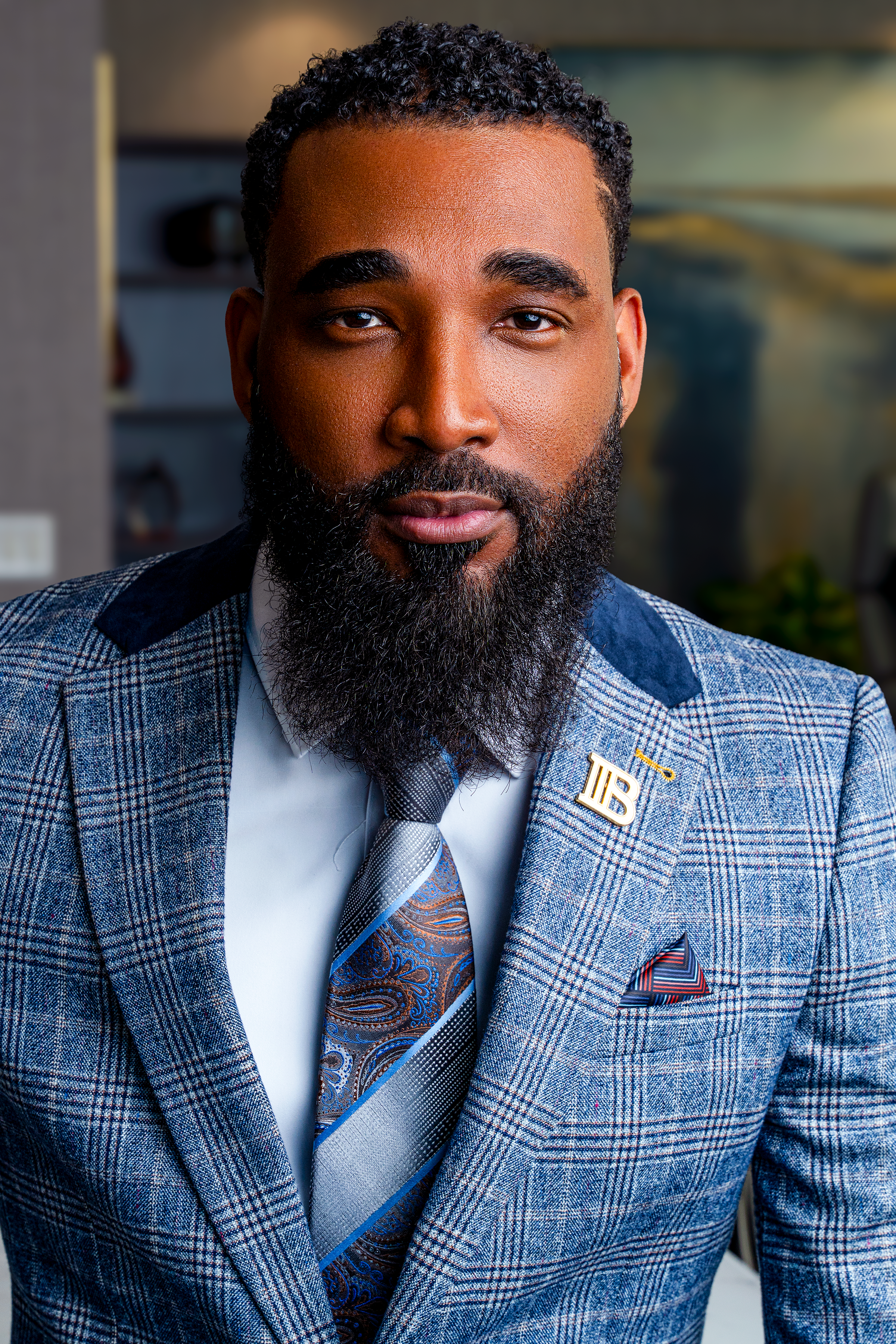 A man with a full beard and mustache wearing a blue plaid suit with a gold lapel pin, white shirt, and patterned tie, looking directly at the camera.