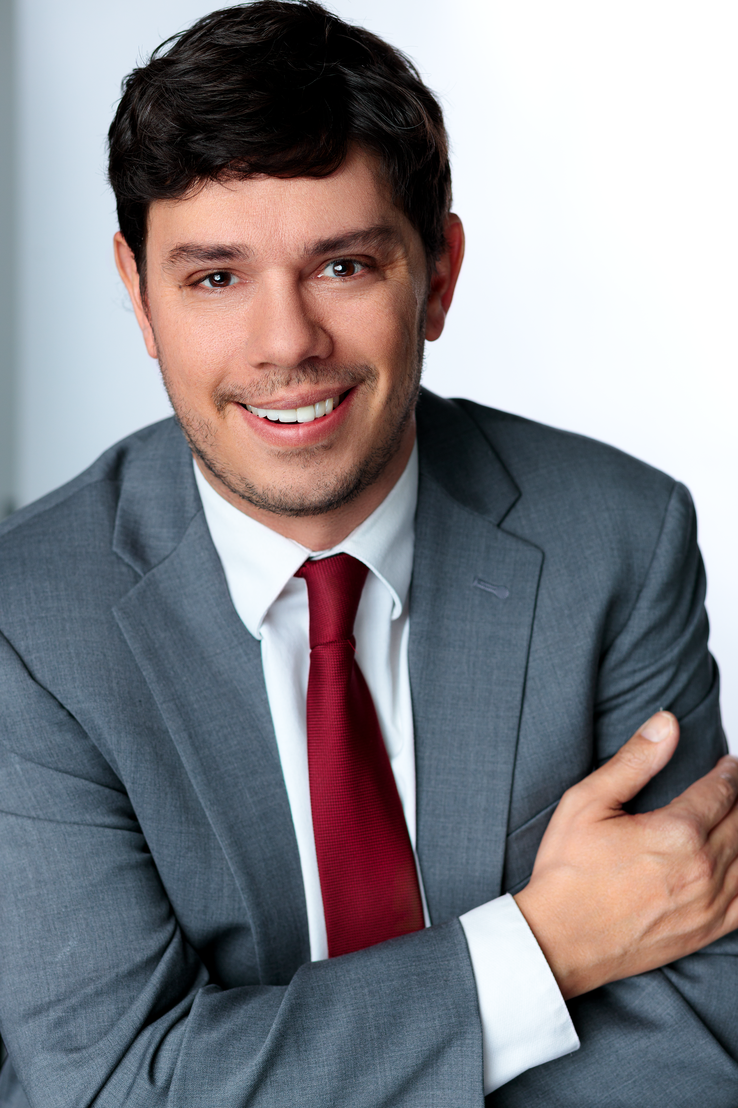 A man in a gray suit with a white shirt and red tie smiling, looking at the camera, with arms crossed in front of a neutral background.