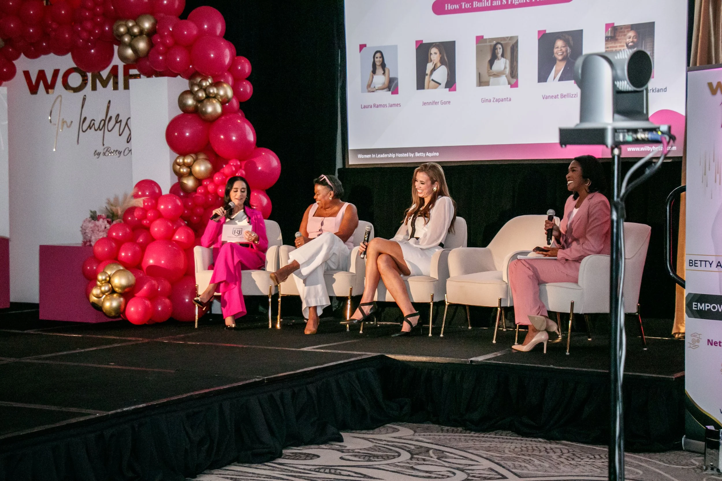 Four women seated on stage at a panel discussion, with a large screen behind them displaying images and names. The stage is decorated with pink and gold balloons.