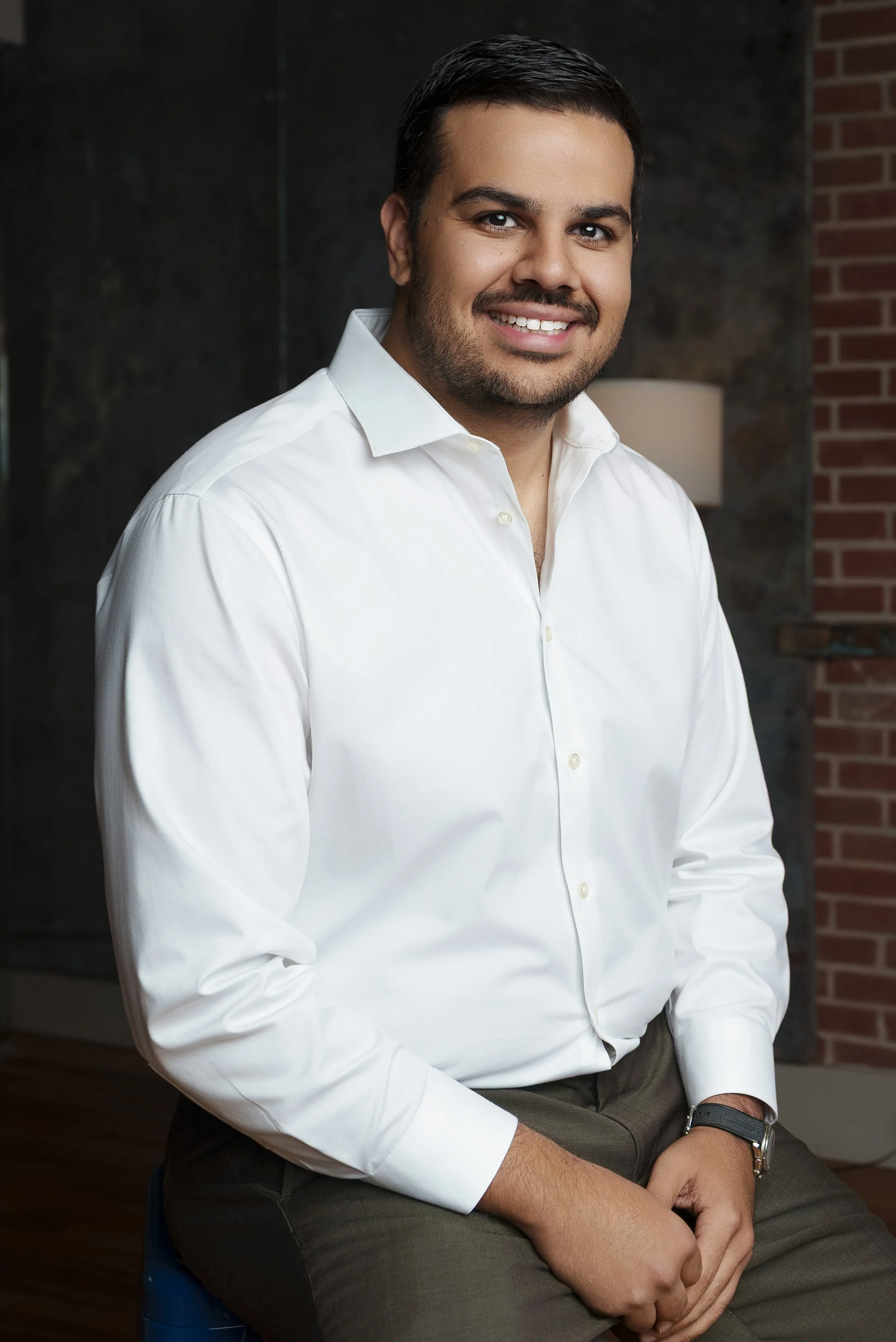 A man with short dark hair, a beard, and a mustache, wearing a white button-up shirt, sitting on a blue stool, smiling in an indoor setting with a dark wall, a lamp, and a brick wall in the background.