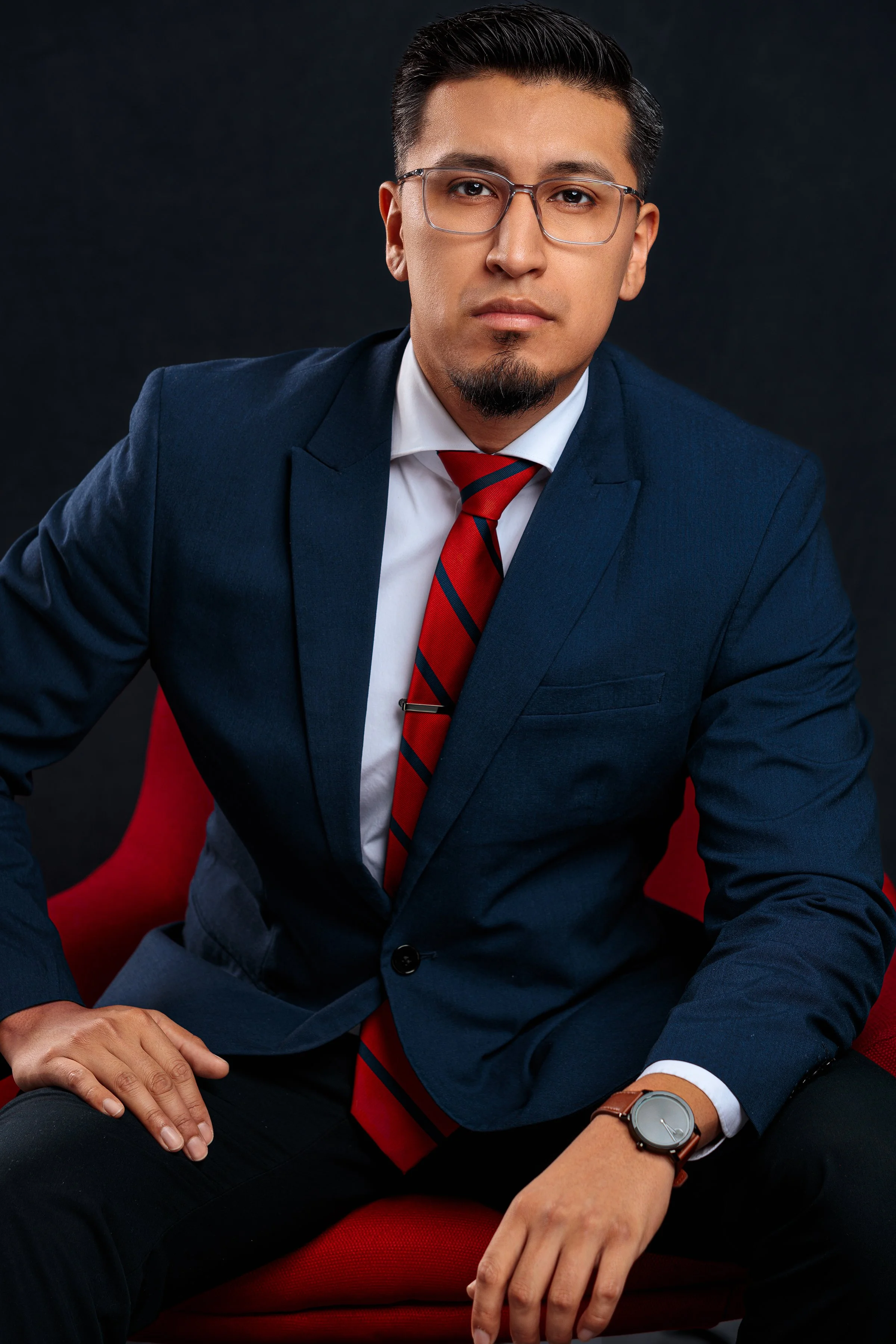 A man wearing glasses, a navy blue suit, a white shirt, and a red and blue striped tie, sitting on a red chair against a dark background.