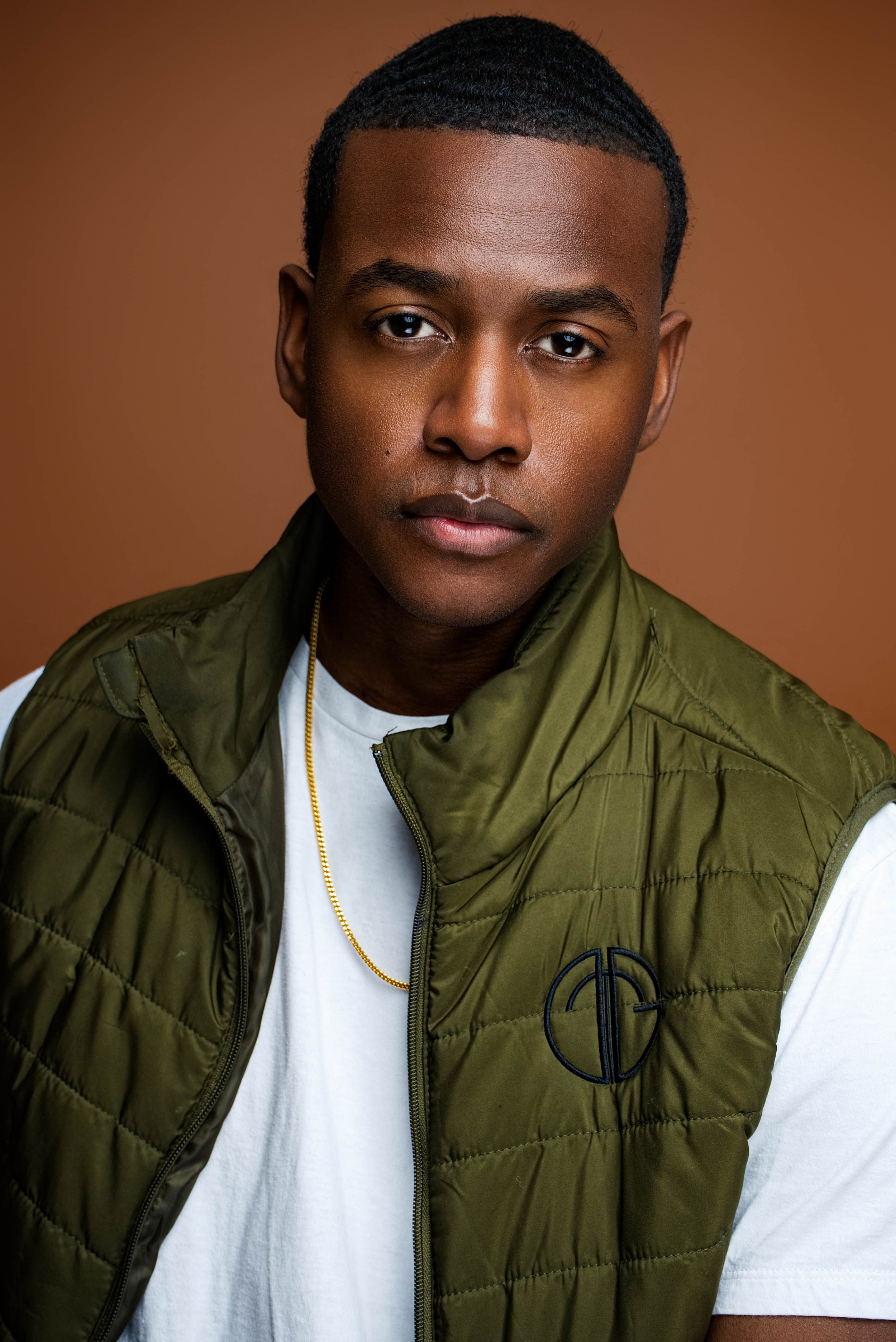 A young man with dark skin and short black hair, wearing a white t-shirt, an olive green padded vest with a logo, and a gold chain, posing against a brown background.