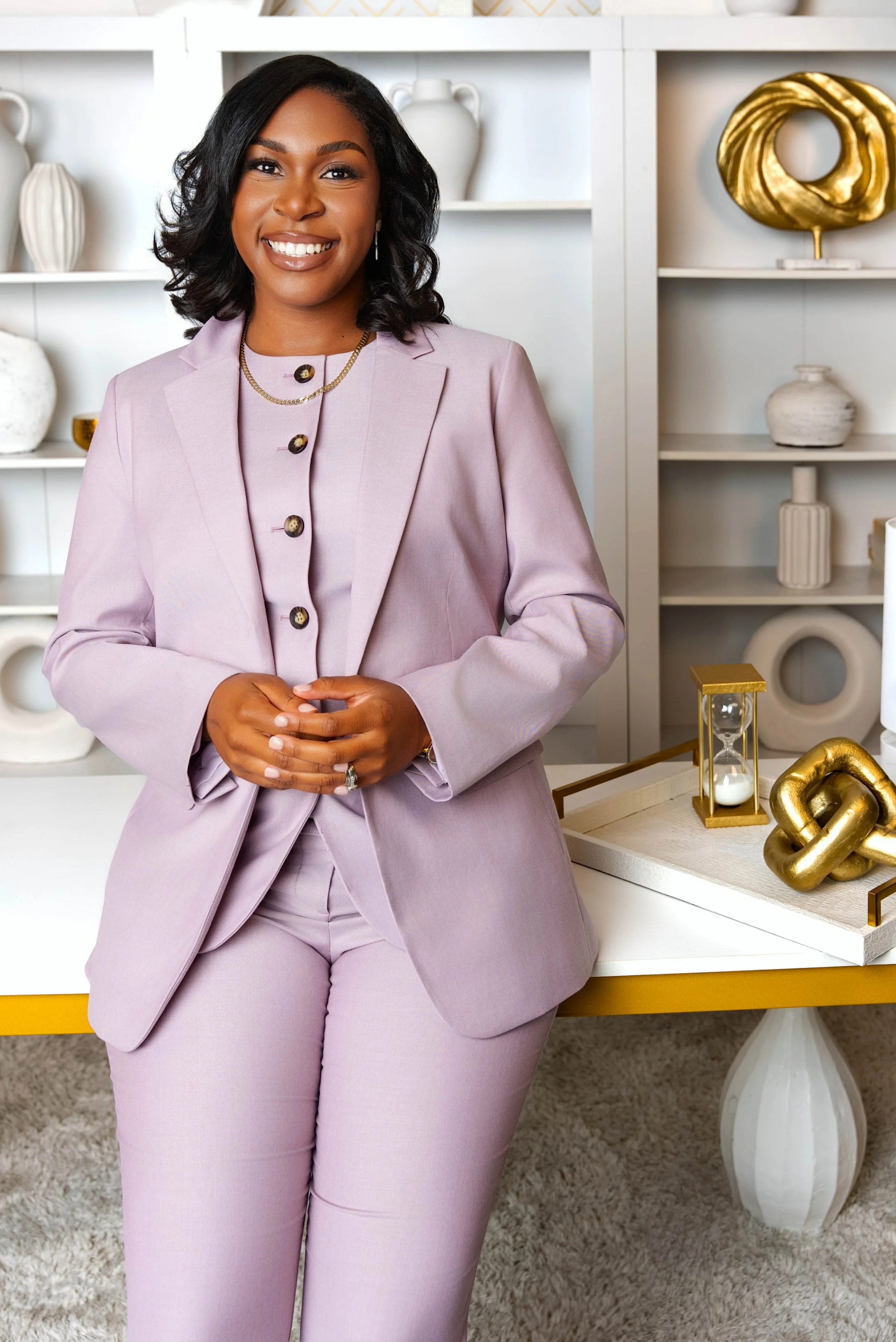 A woman in a lavender suit standing in front of white shelves decorated with white and gold vases, with a white and gold decorative sculpture on a tray in front.