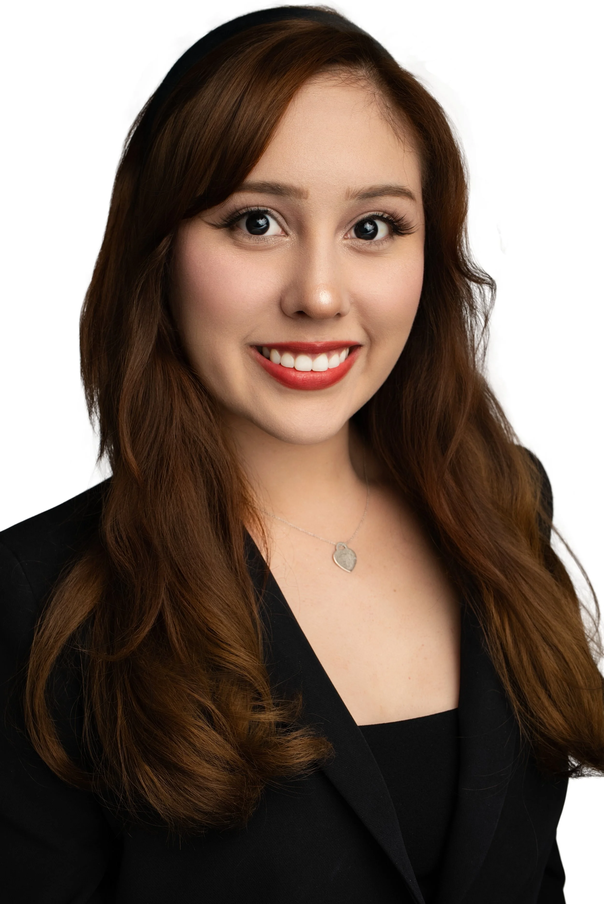 A woman with long, wavy brown hair, smiling showing white teeth, wearing a black blazer, a silver heart-shaped necklace, and a black headband, on a plain white background.