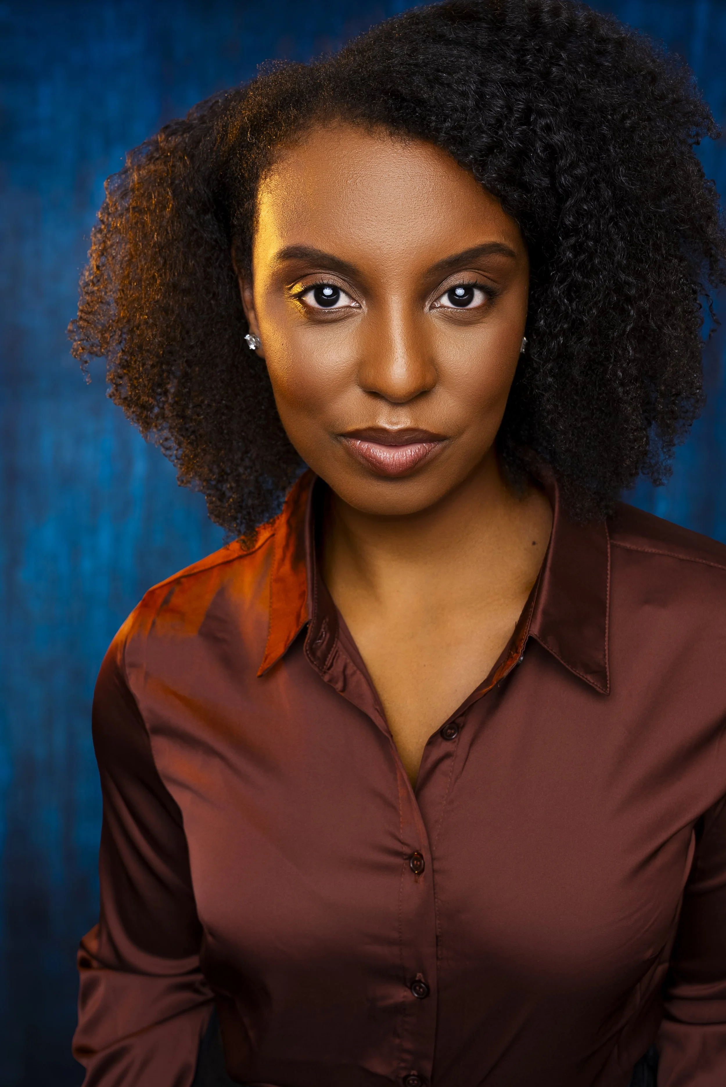 Portrait of a woman with dark, curly hair, wearing a satin brown blouse, standing in front of a blue curtain background.