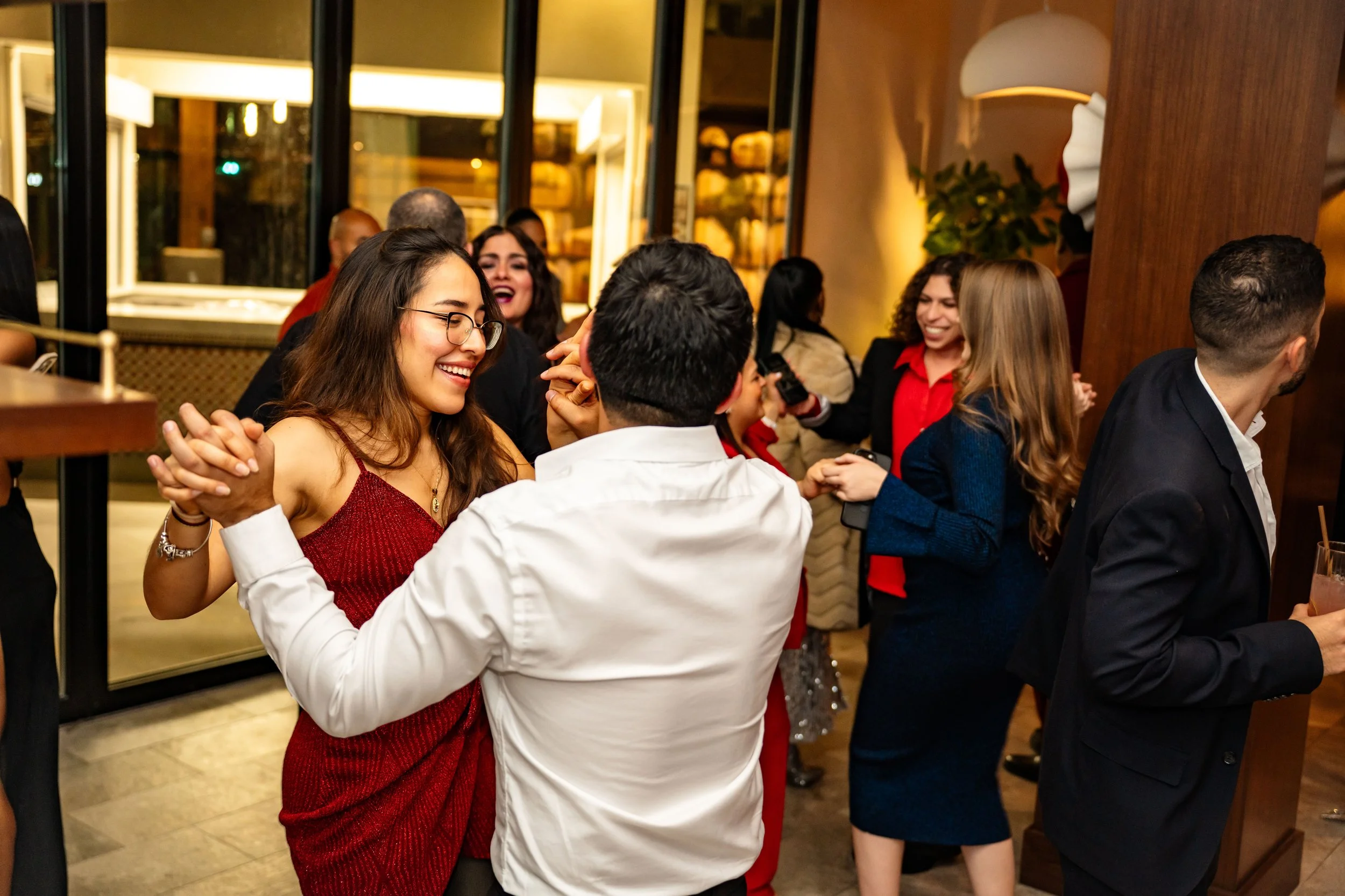A young woman in a red dress dancing with a man in a white shirt at a lively social gathering, surrounded by other people chatting and enjoying themselves.