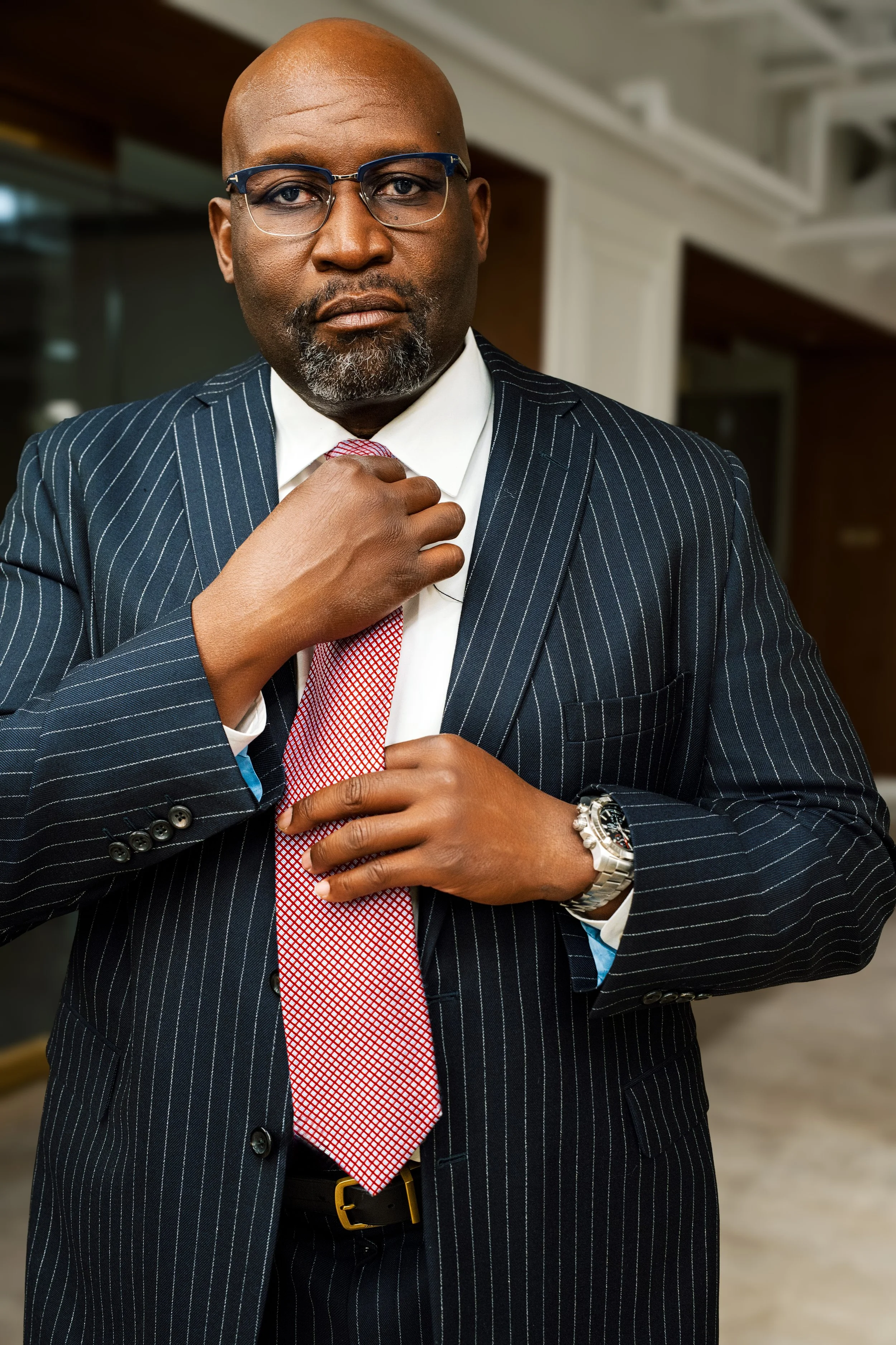 A suited man adjusting his tie. He is wearing glasses, a wristwatch, and a pinstriped navy suit.