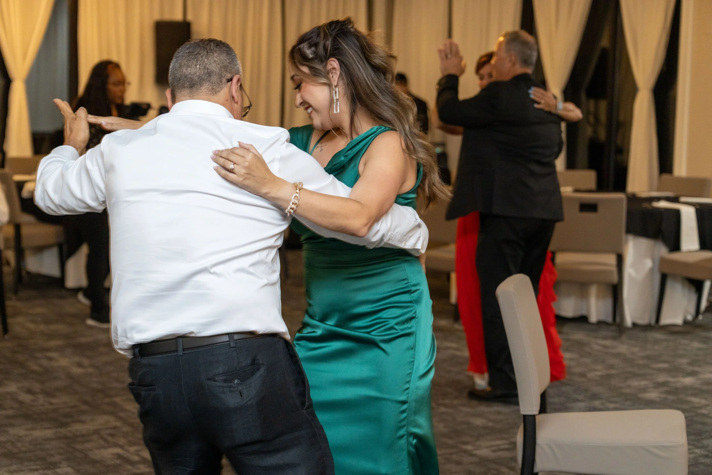 People dancing at a social event in a banquet hall, with a woman in a teal dress and a man in a white shirt in the foreground.