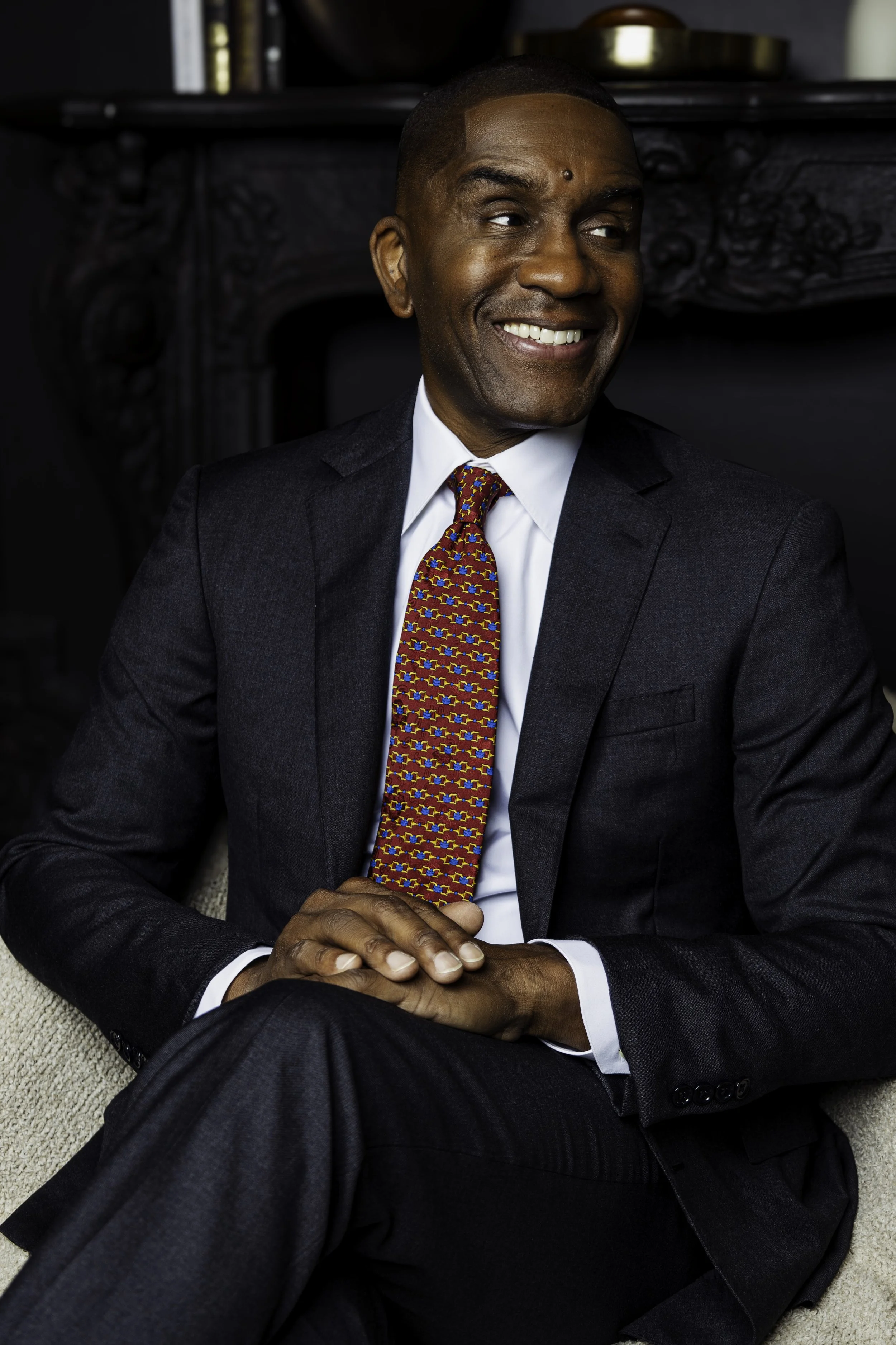 A smiling man in a dark suit with a colorful tie, sitting with hands clasped on his lap in front of a dark fireplace.