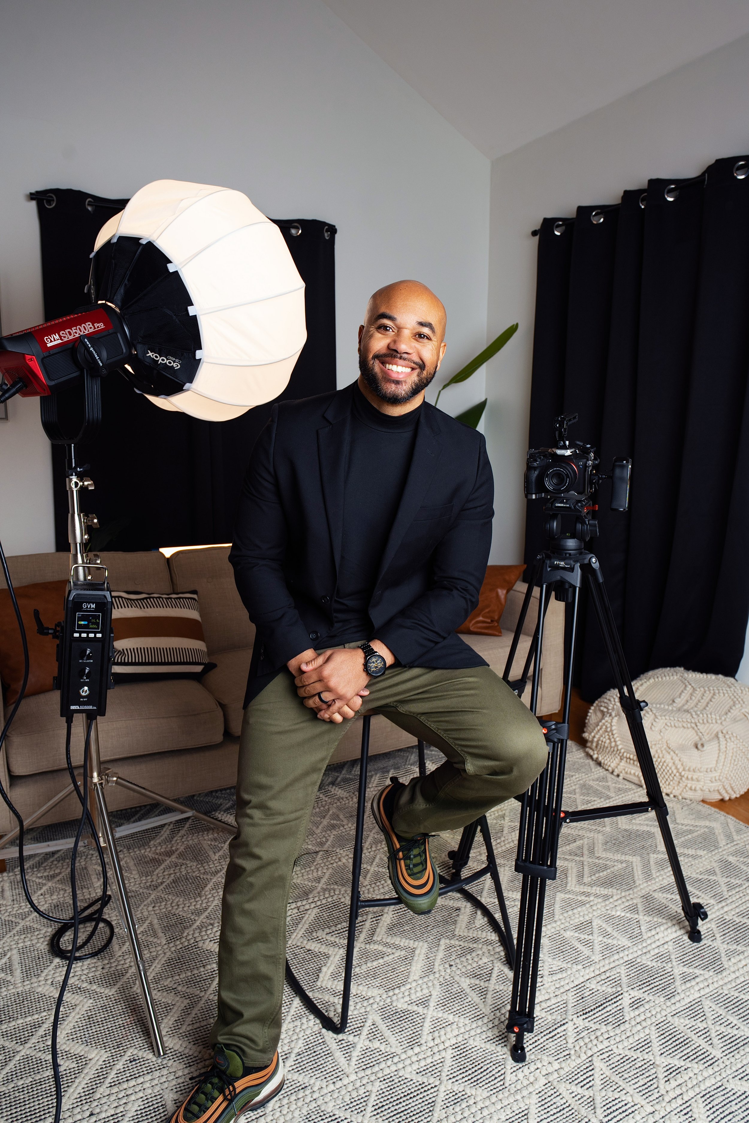 A man in a black blazer and khaki pants sitting on a stool in a home studio with professional lighting and a camera on a tripod, smiling at the camera.