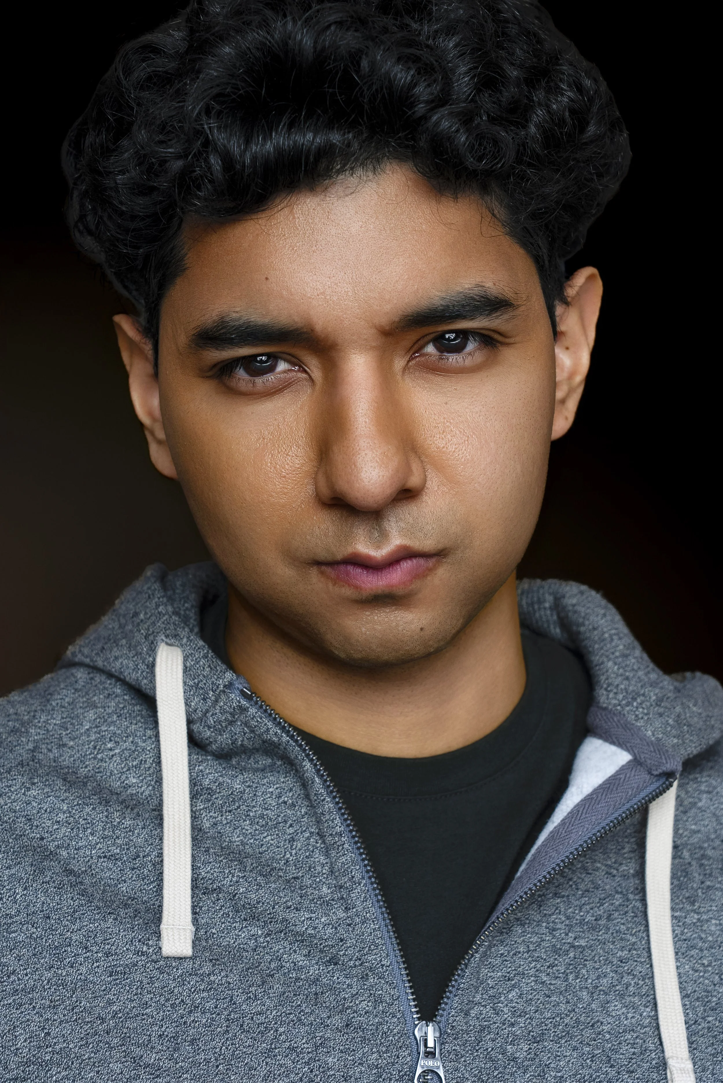 Close-up portrait of a young man with dark curly hair wearing a gray zip-up hoodie and black shirt against a dark background.