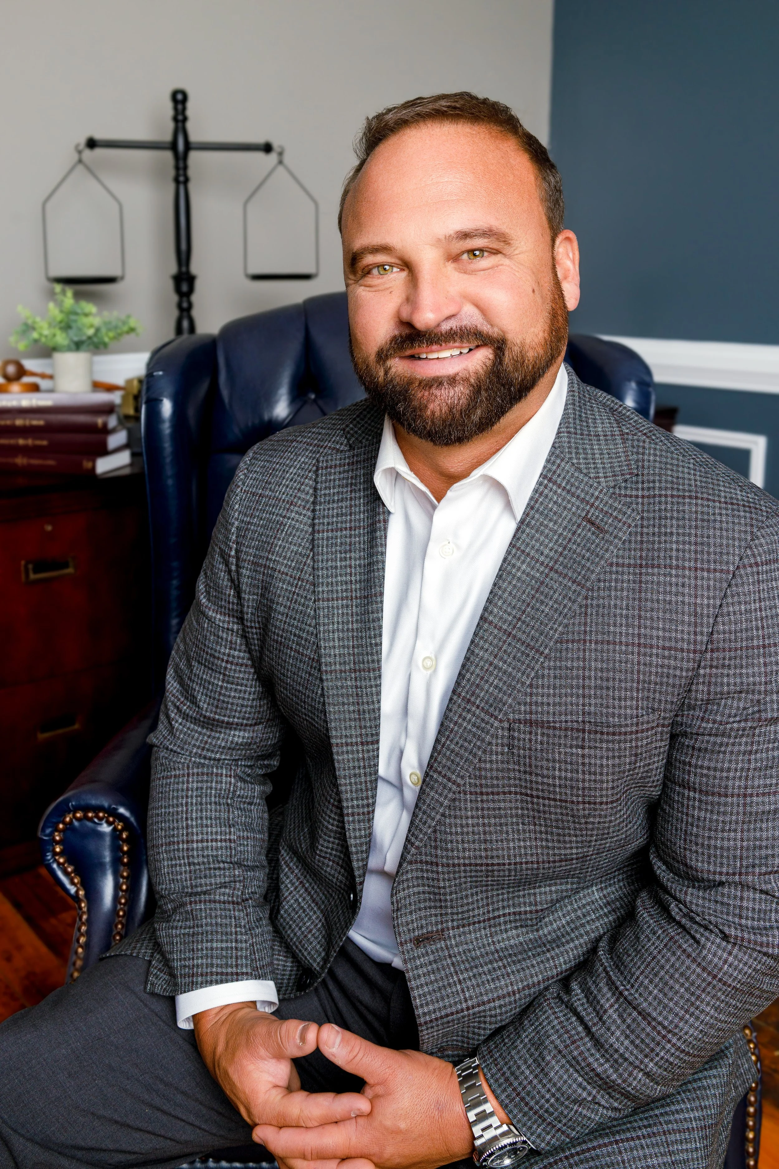 A smiling man wearing a gray checkered suit jacket and white shirt, sitting in a leather office chair in a professional office setting.