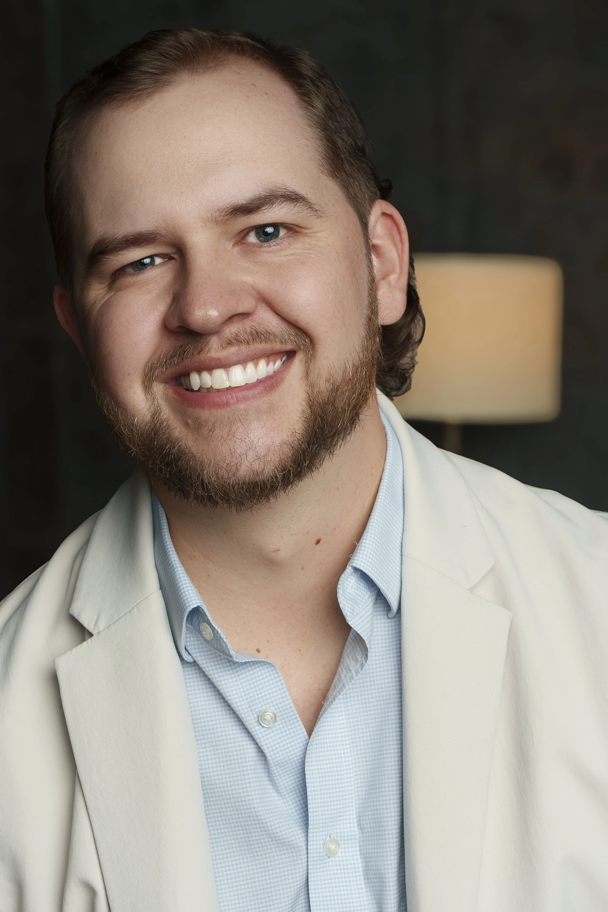 Close-up portrait of a smiling man with a beard, wearing a light-colored blazer and blue shirt, indoors with a blurred background and a lamp.