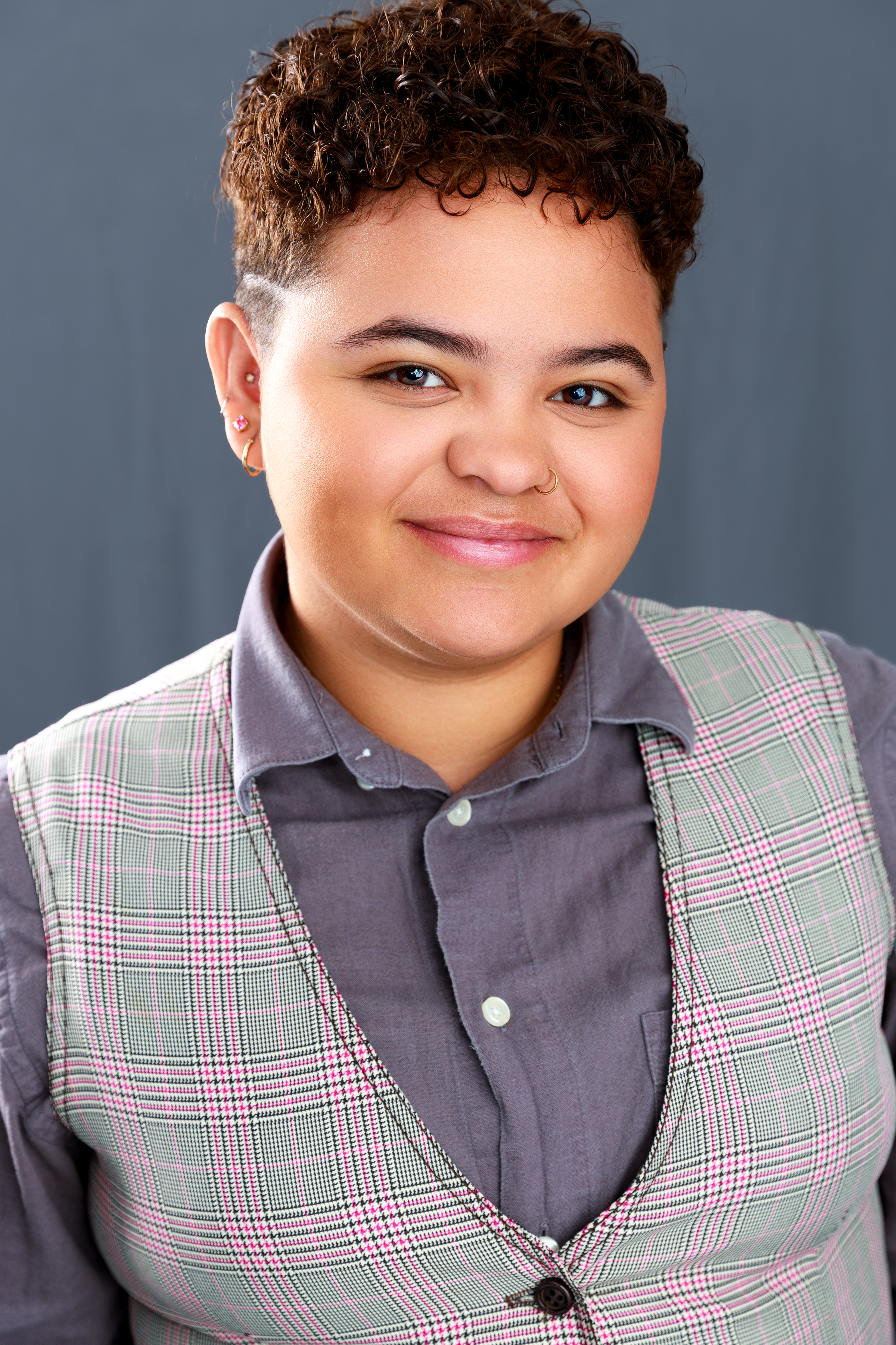 A young woman with short curly hair and multiple earrings, wearing a gray button-up shirt and a plaid vest, smiling at the camera against a plain gray background.