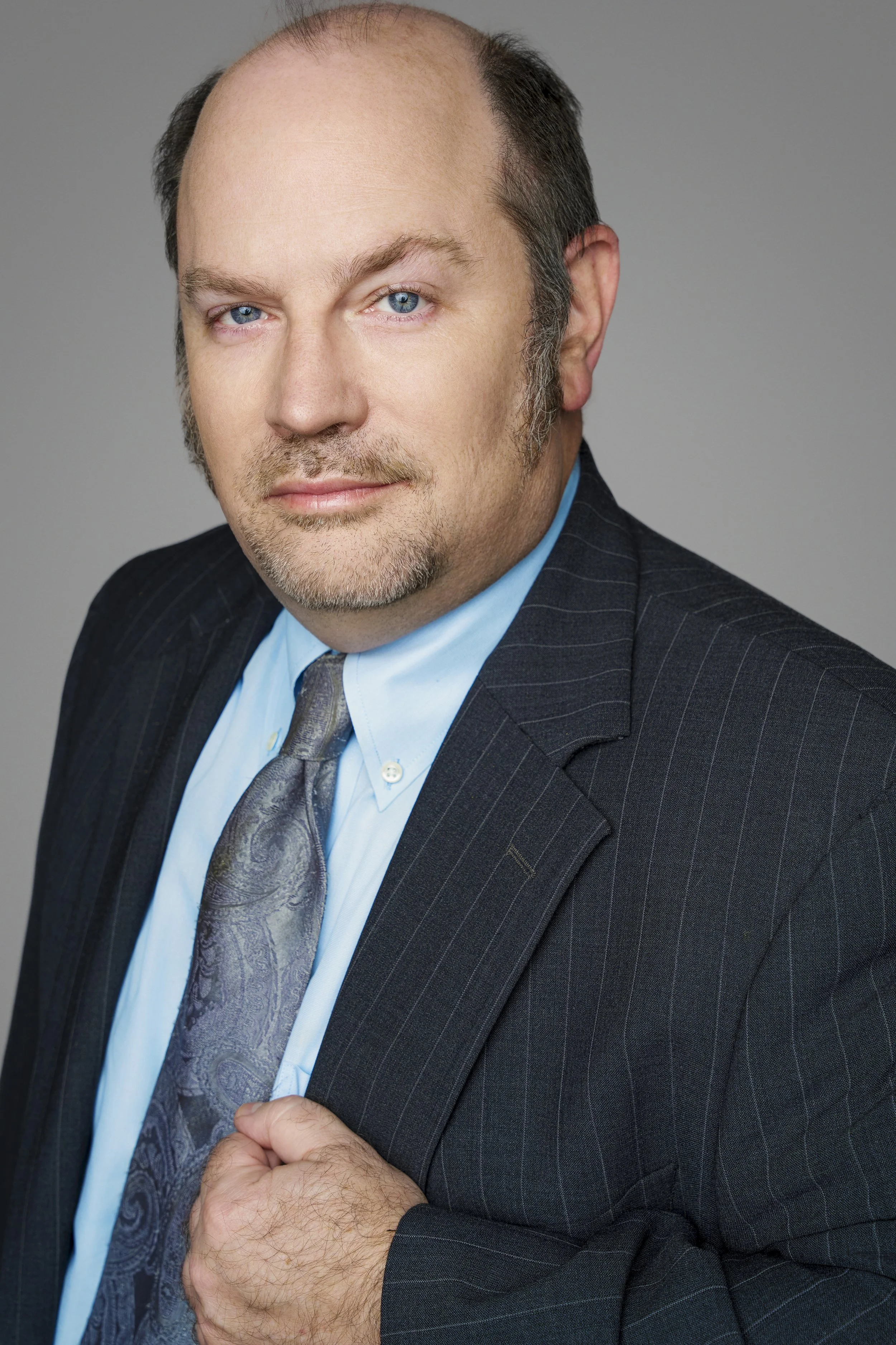 A middle-aged man with light skin, blue eyes, and a goatee, dressed in a dark pinstripe suit, light blue shirt, and a patterned tie, looking into the camera with a neutral expression.