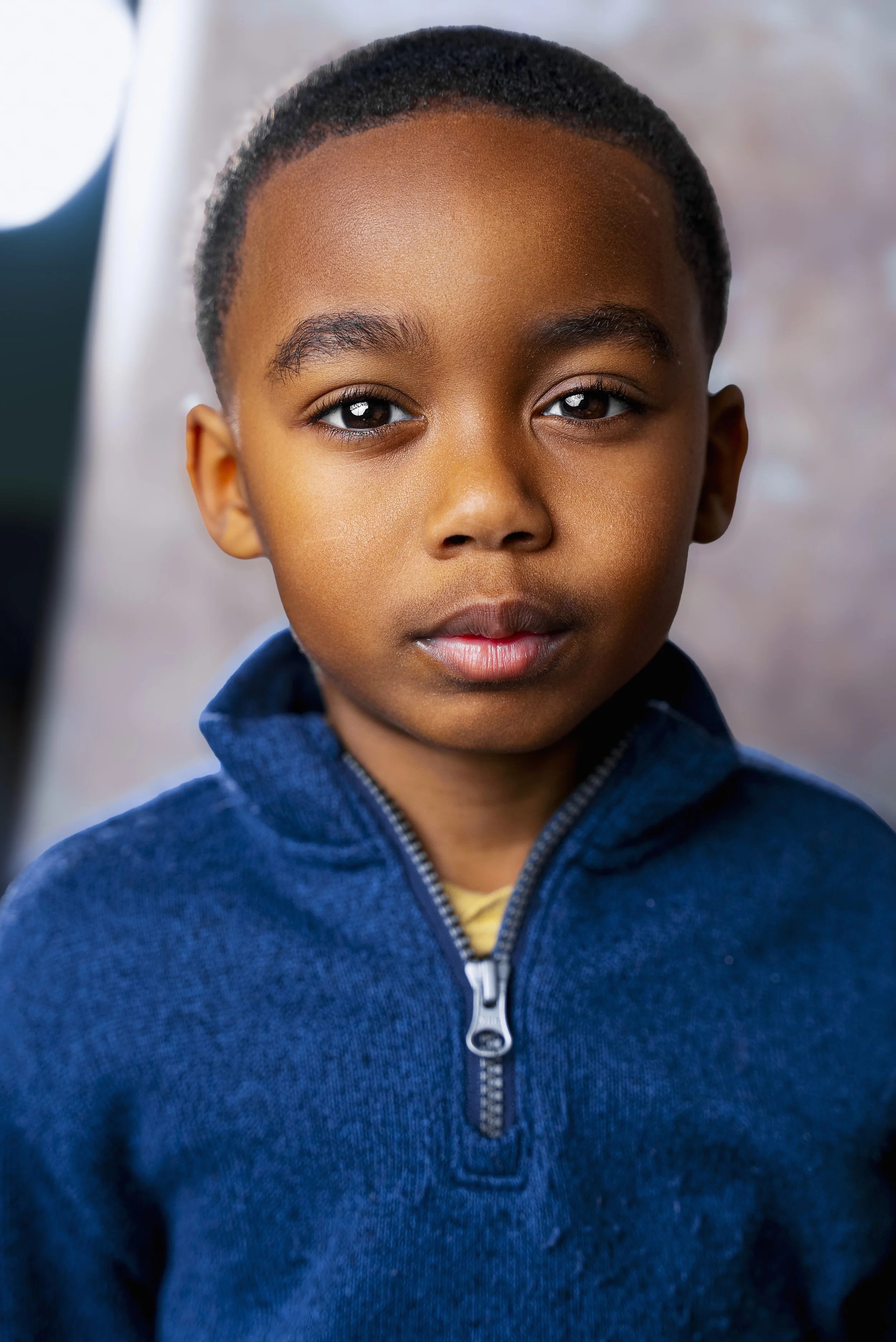 A young boy with brown skin and short curly hair, wearing a blue zip-up sweater, looking directly at the camera with a neutral expression.