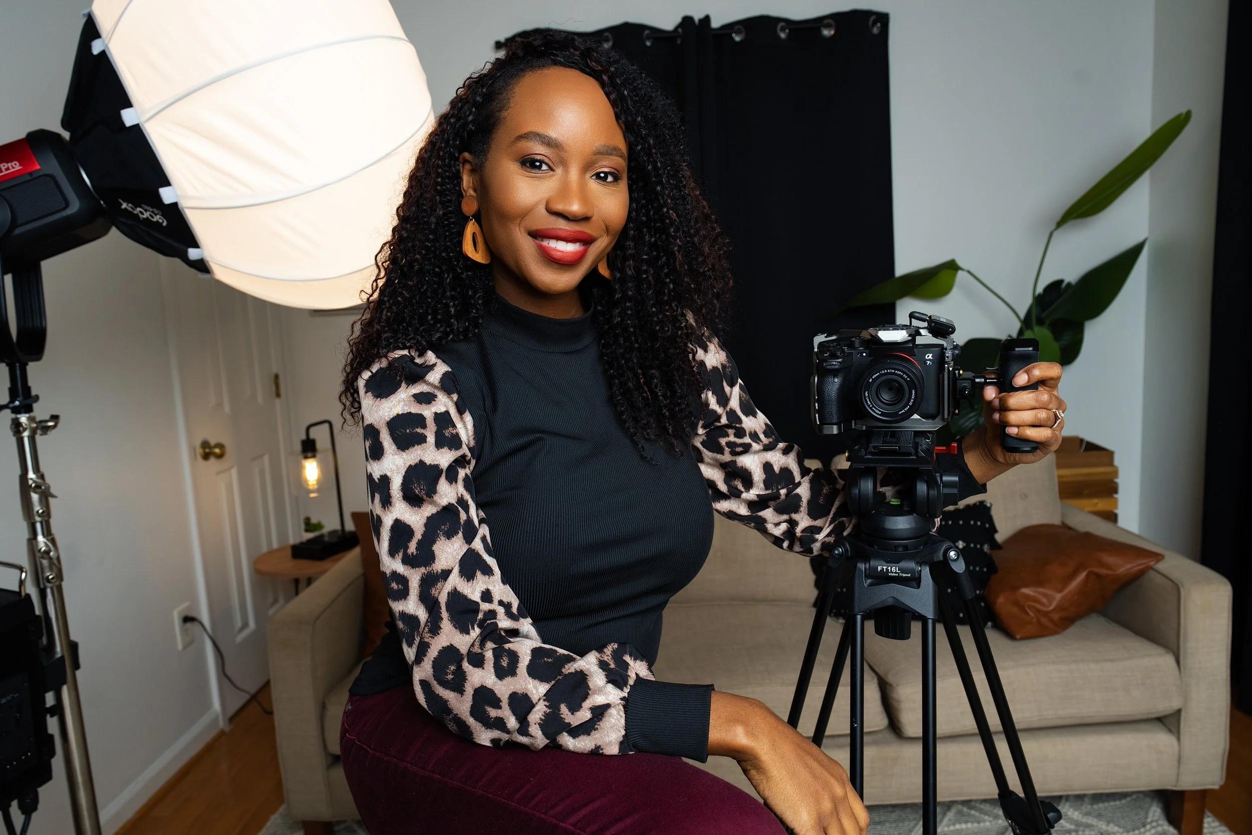 A woman with curly hair, wearing a black top with leopard print sleeves, sits in a room with a camera on a tripod, professional lighting, a beige couch, and a black curtain in the background.