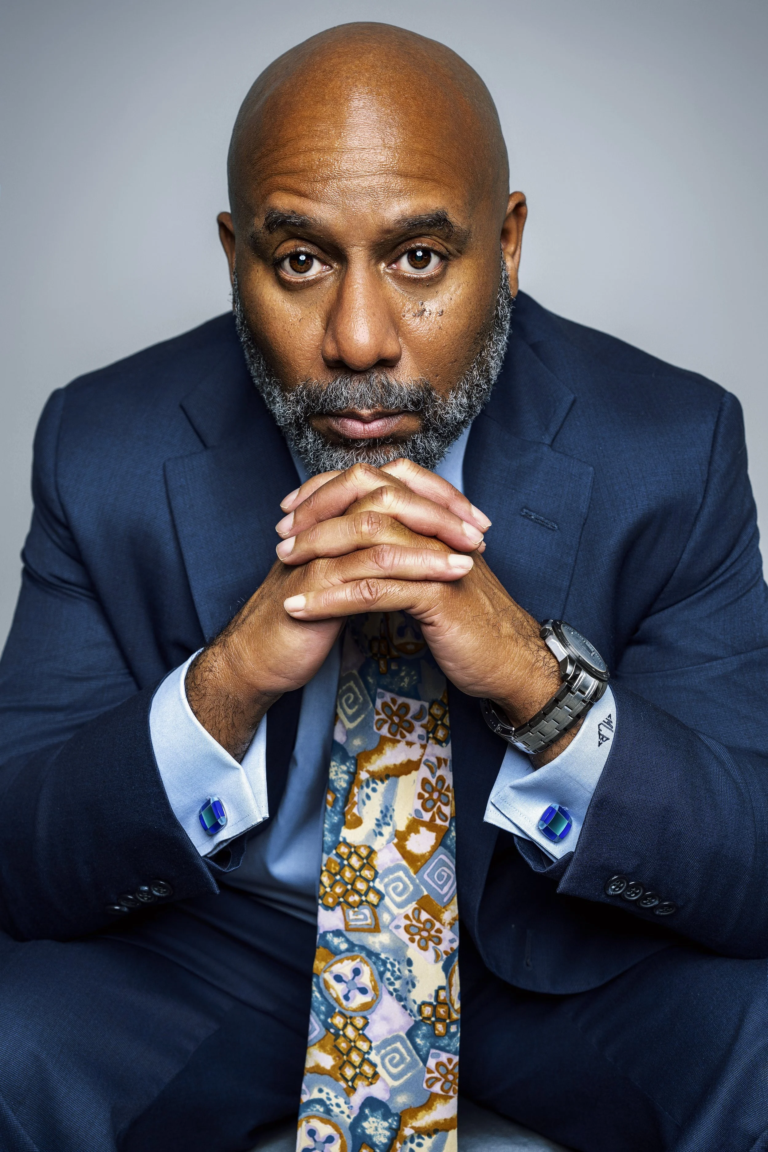 A man wearing a navy blue suit, light blue shirt, and a colorful patterned tie, with a beard and a bald head, looking directly at the camera with hands clasped in front of his face.