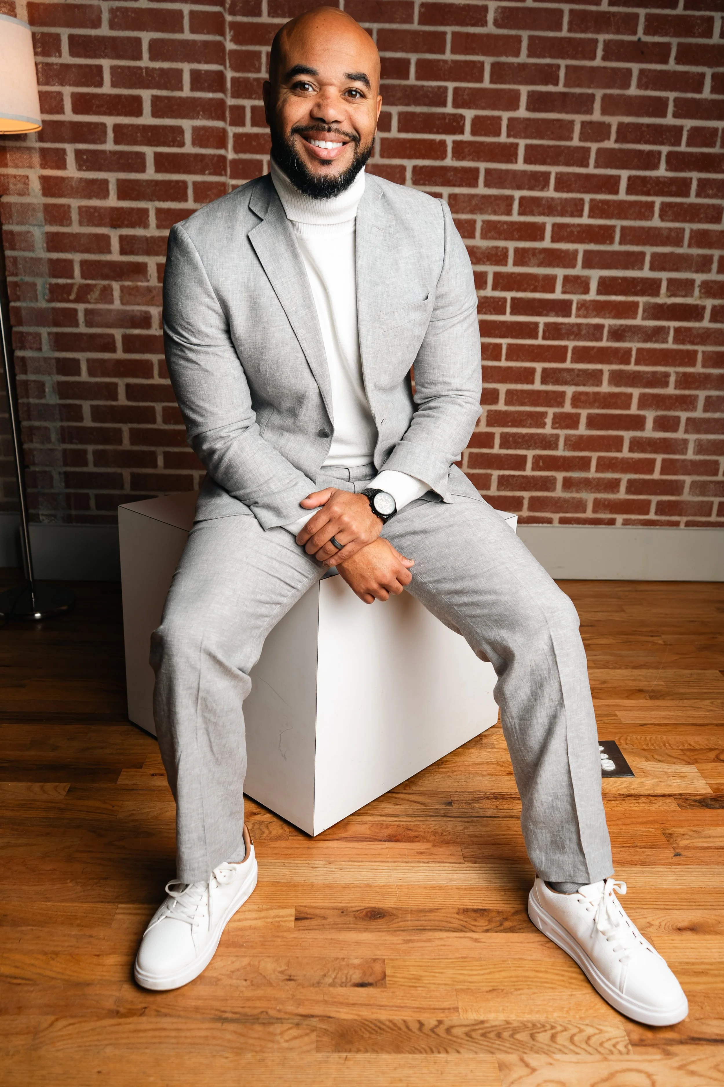 A man sitting on a white cube, wearing a light gray suit, white turtleneck, and white sneakers, smiling in front of a brick wall.