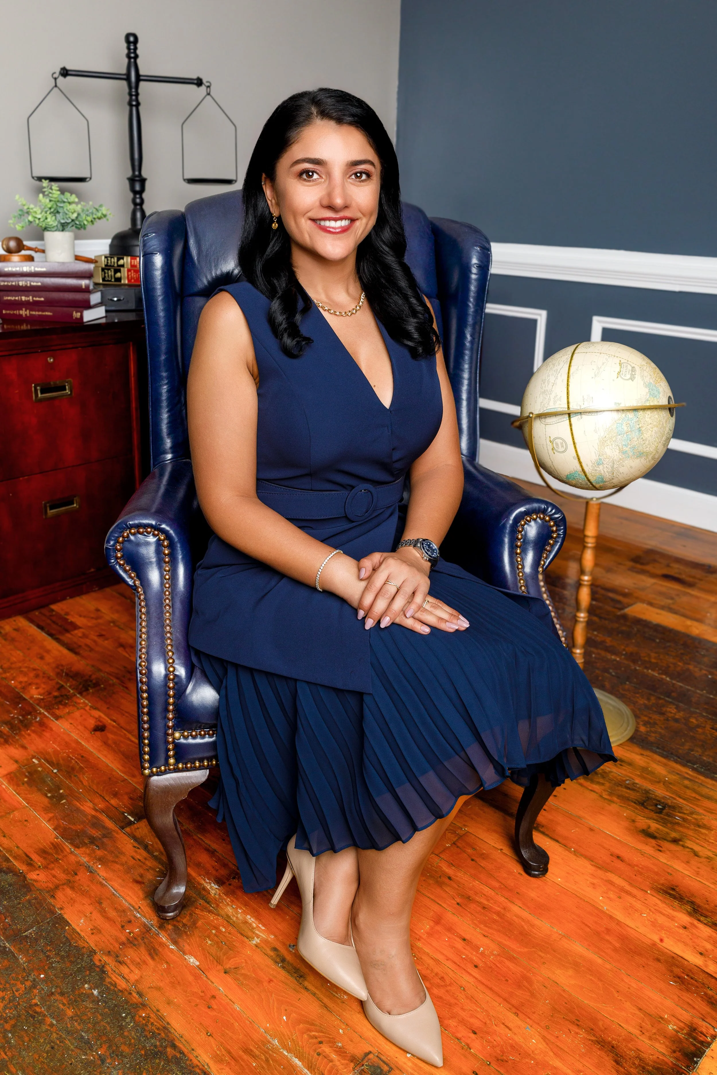 A woman in a navy blue dress sitting on a leather armchair in an office setting with a globe and books nearby.