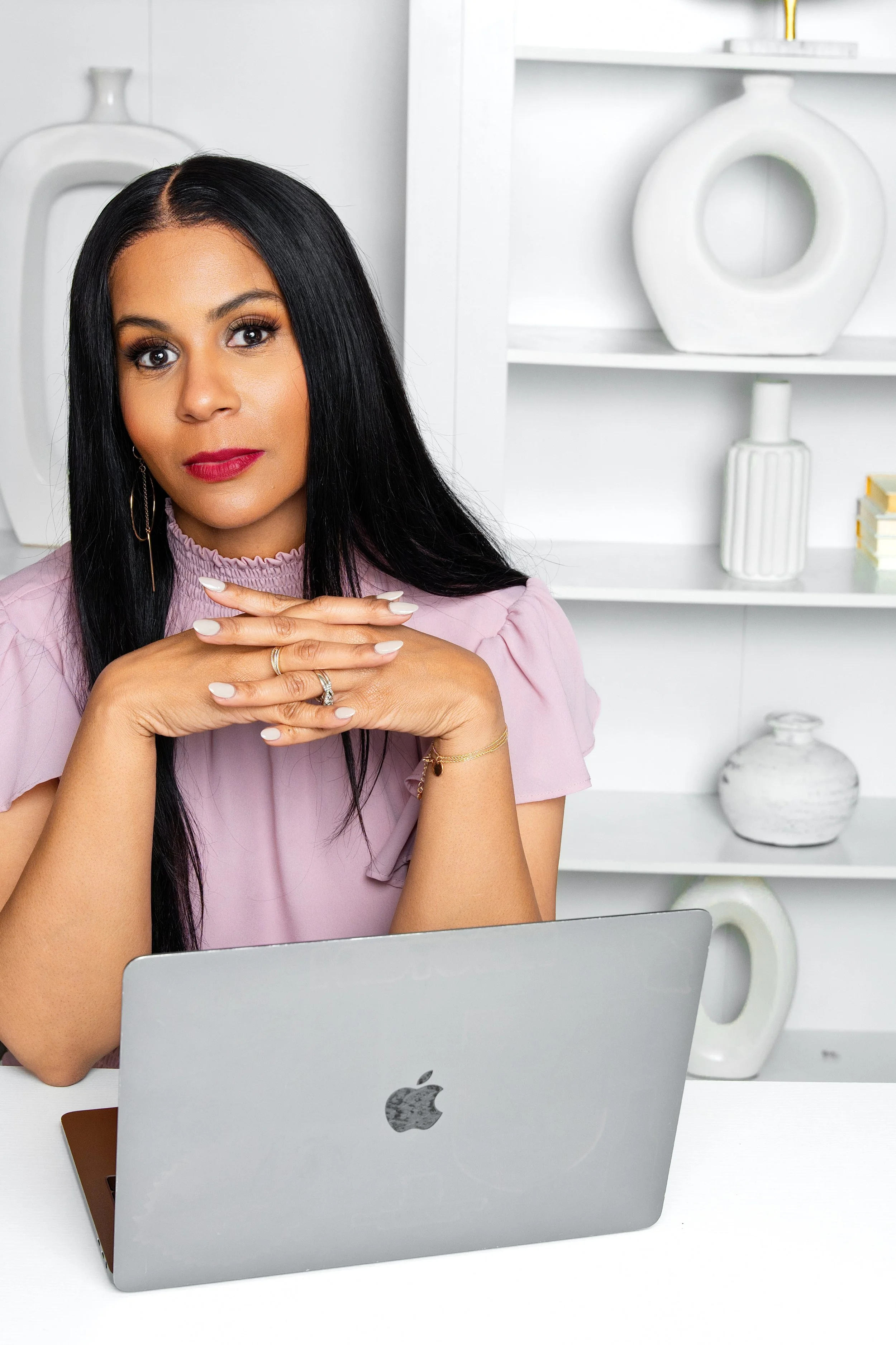 A woman with long black hair, wearing a light purple top and jewelry, sitting at a white desk with a silver MacBook in front of her, in a bright room with white shelves and decorative vases in the background.