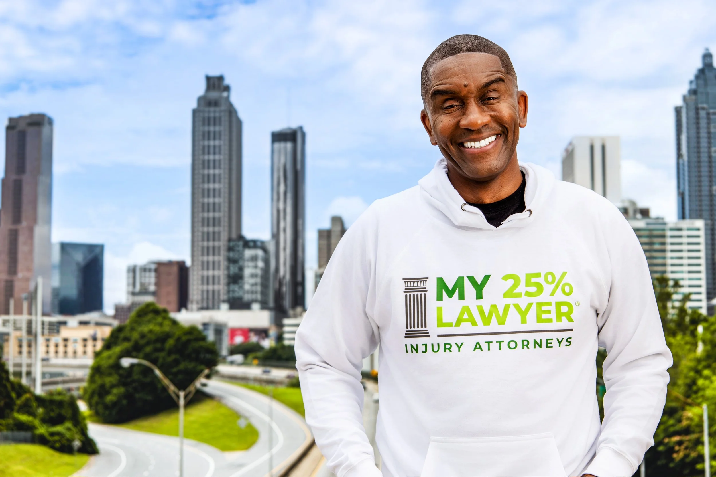 A man smiling outdoors in front of a city skyline wearing a white hoodie with green and black text that reads 'My 25% Lawyer Injury Attorneys'.