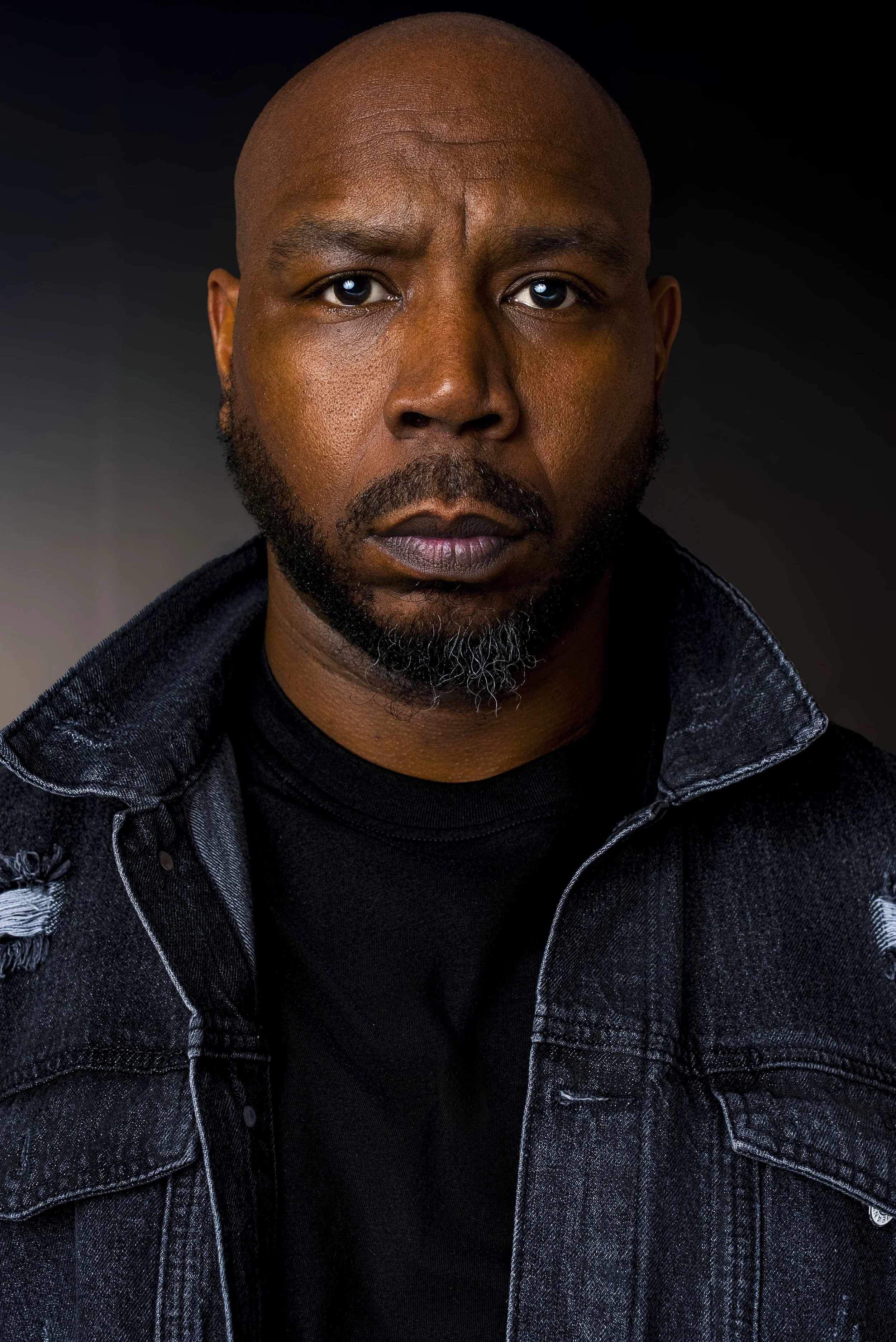 A close-up portrait of a bald Black man with a beard, wearing a black shirt and a distressed black denim jacket, against a dark background.