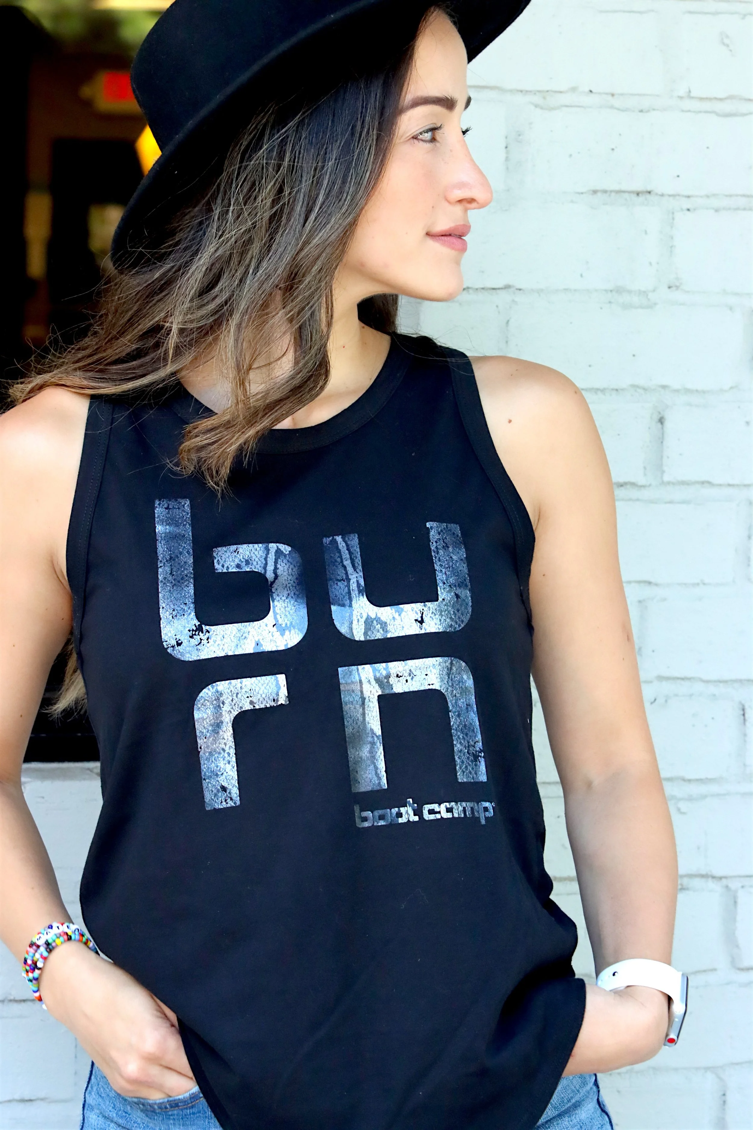 Young woman with brown hair in a black tank top with a logo, wearing a black hat and a white fitness tracker, standing against a white brick wall.