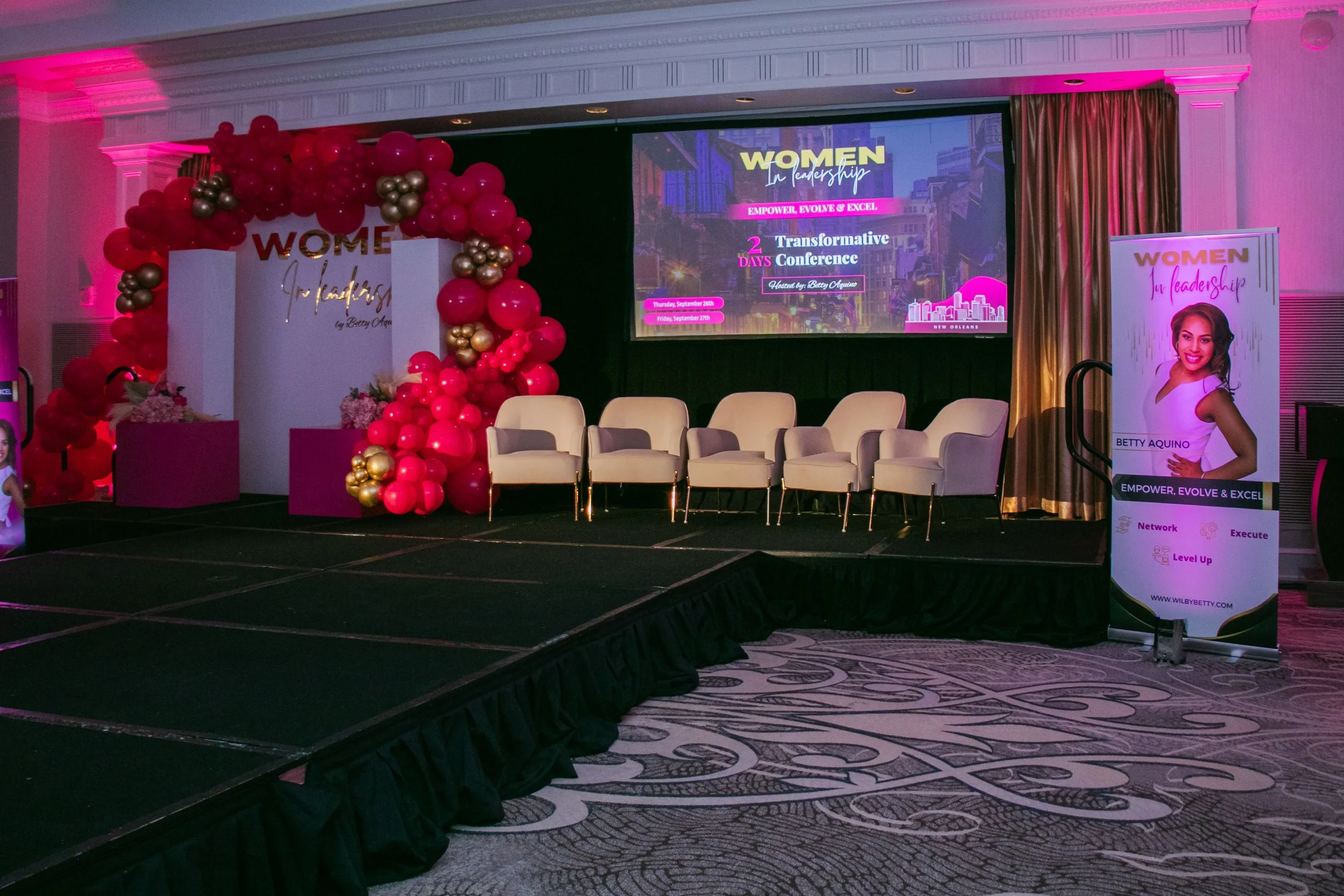 Stage setup for a women's leadership conference with a balloon arch, chairs, and a digital screen displaying conference details.