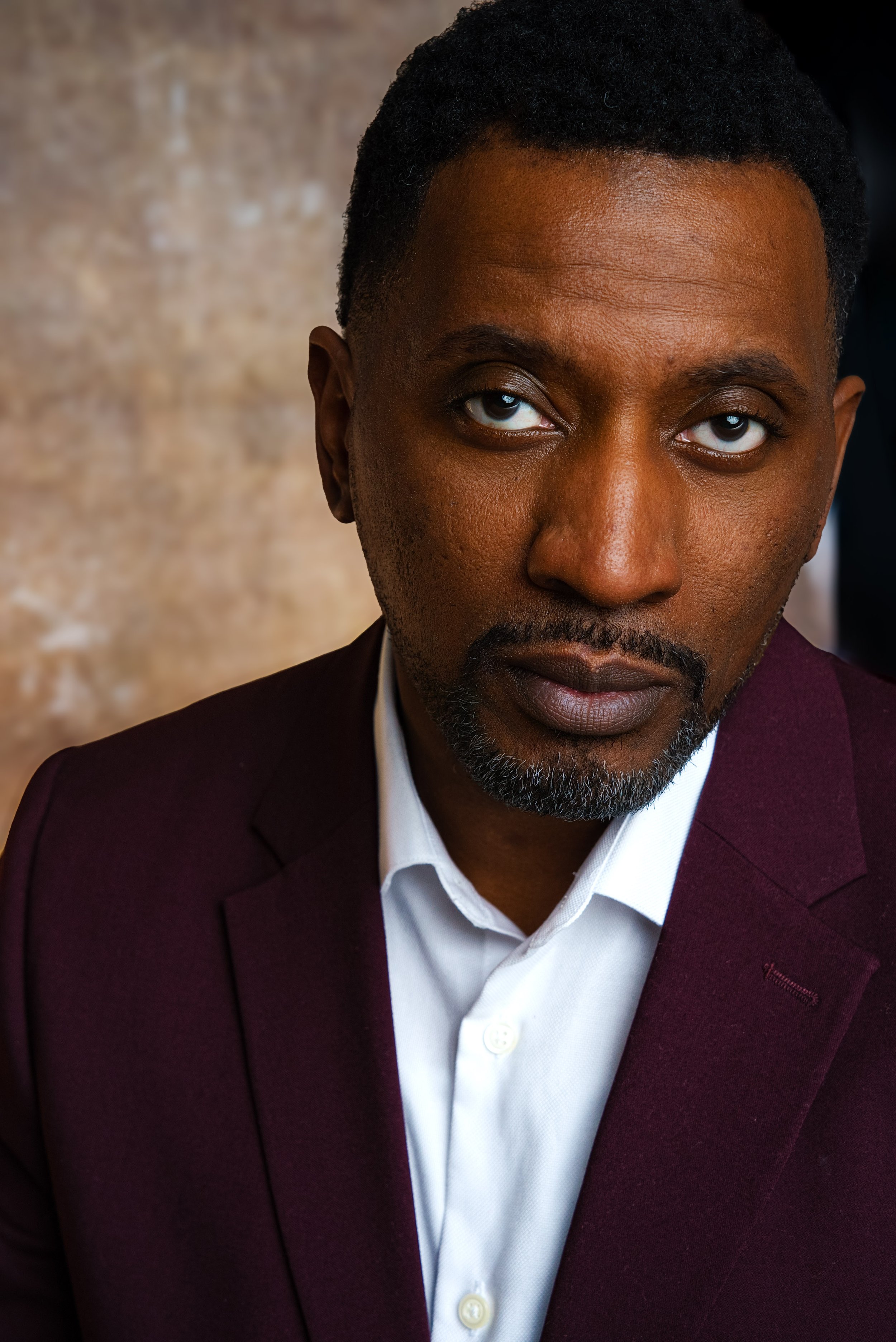Close-up of a man wearing a dark maroon blazer and white shirt, looking intensely at the camera.