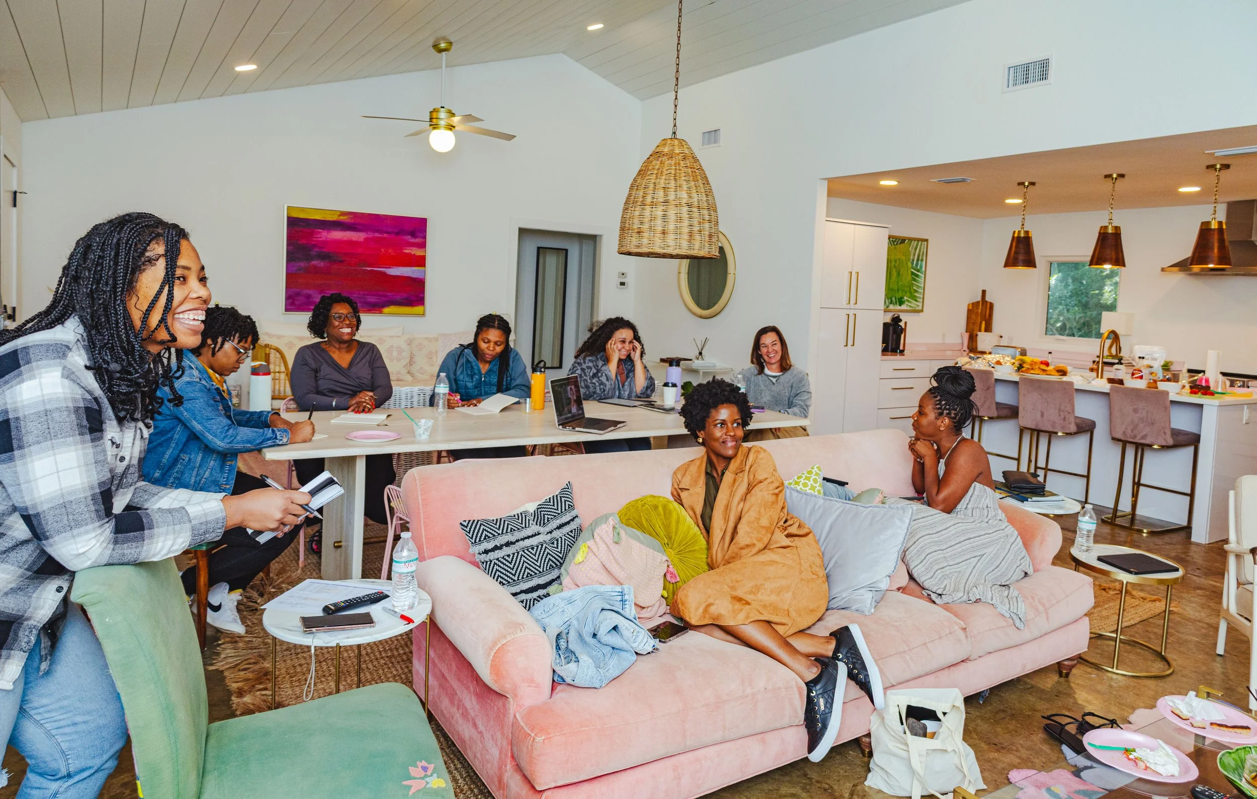 Group of women socializing in a bright, modern living room with an open kitchen. Some women sit on a pink sofa, while others stand or sit at the dining table. The room has colorful artwork, patterned pillows, and various drinks and snacks on the tabl