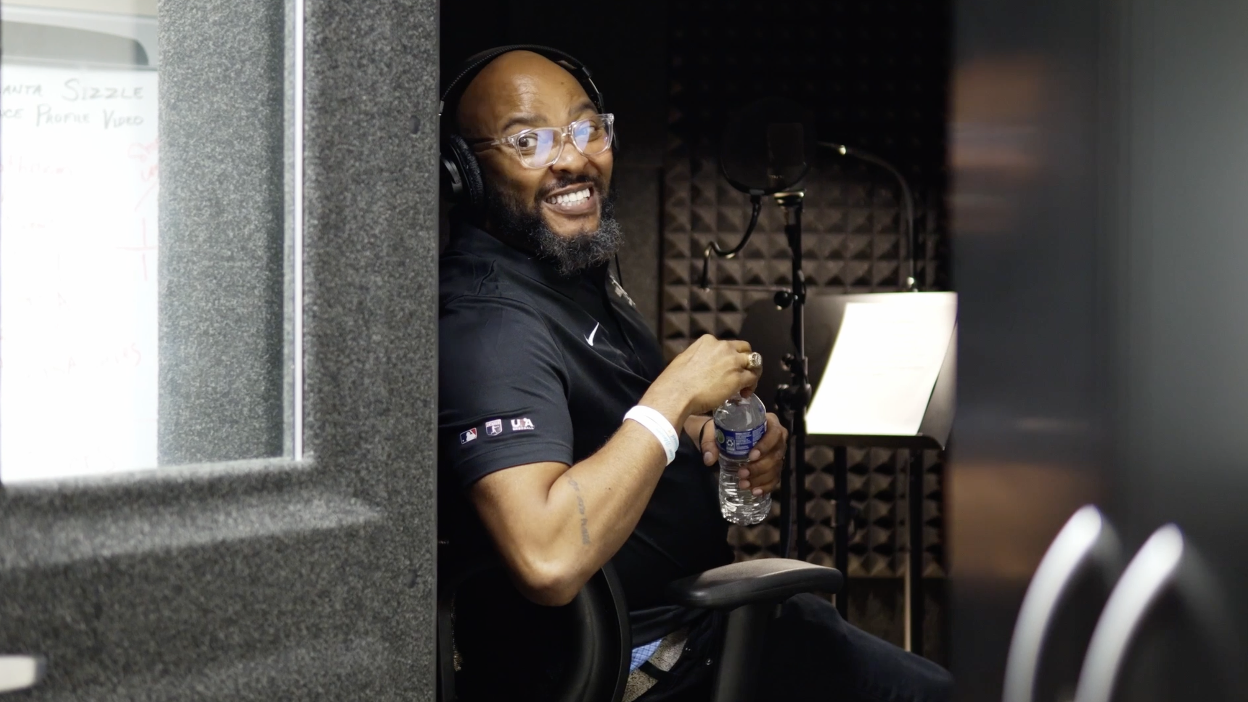 A man with glasses, headphones, and a beard, sitting in a recording studio, smiling, holding a water bottle.
