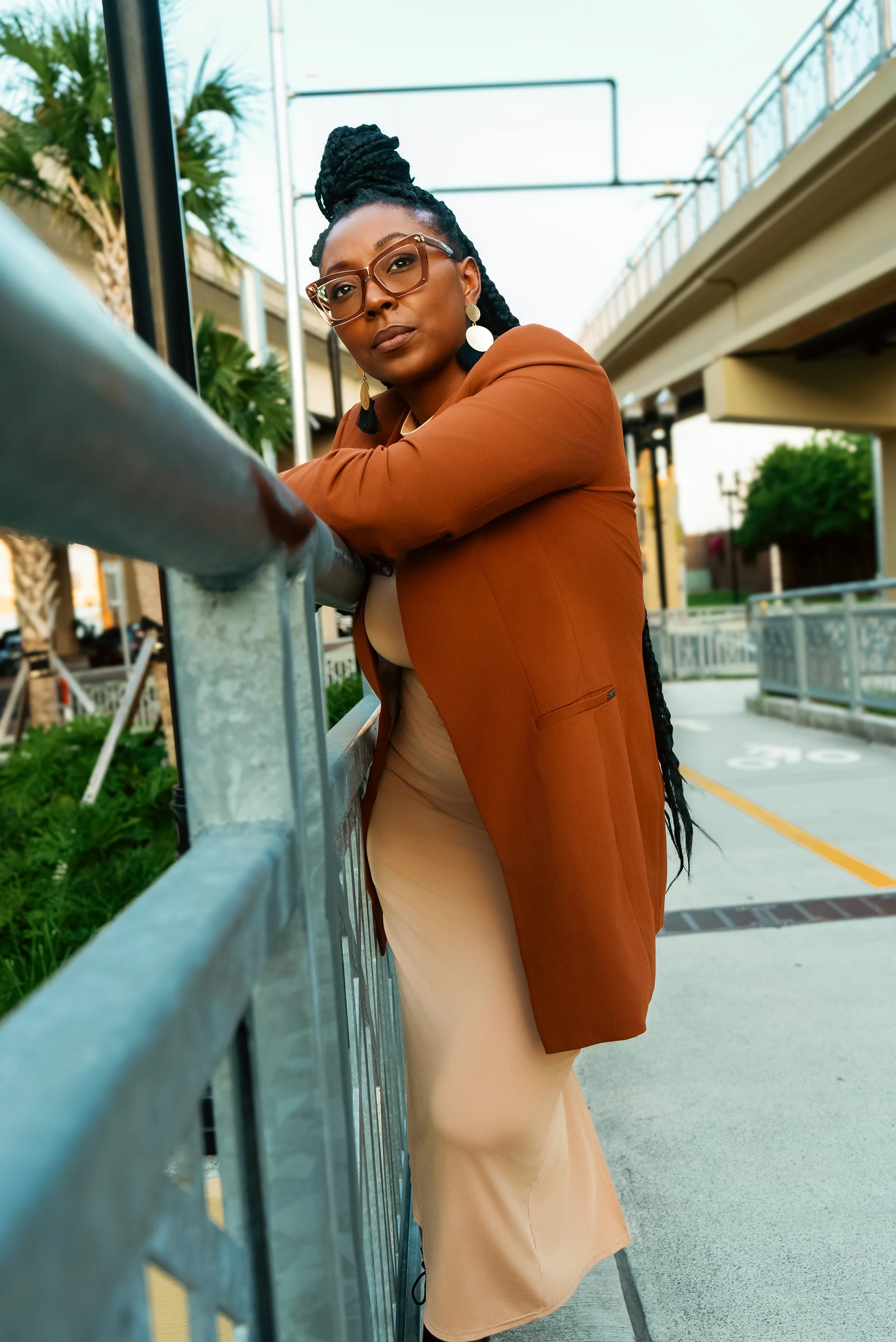 A woman with glasses and hoop earrings leaning on a metal railing outdoors, wearing a tan dress and a brown blazer, with buildings and trees in the background.