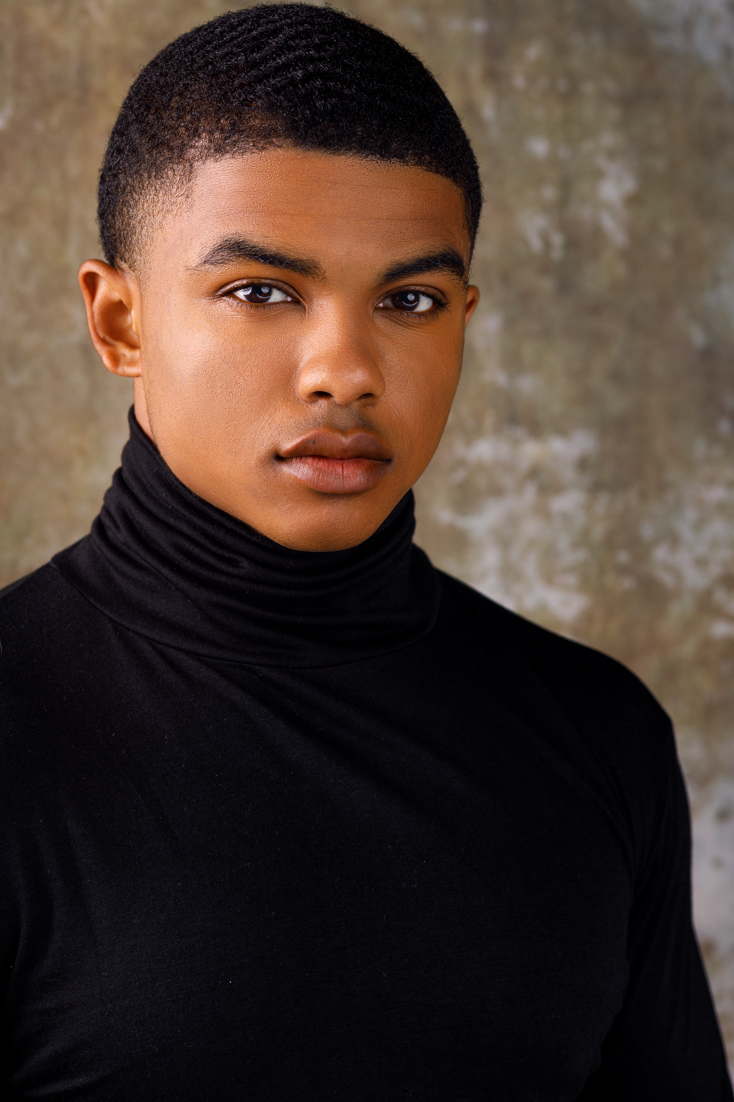 Portrait of a young person with short hair, wearing a black turtleneck, against a textured background.