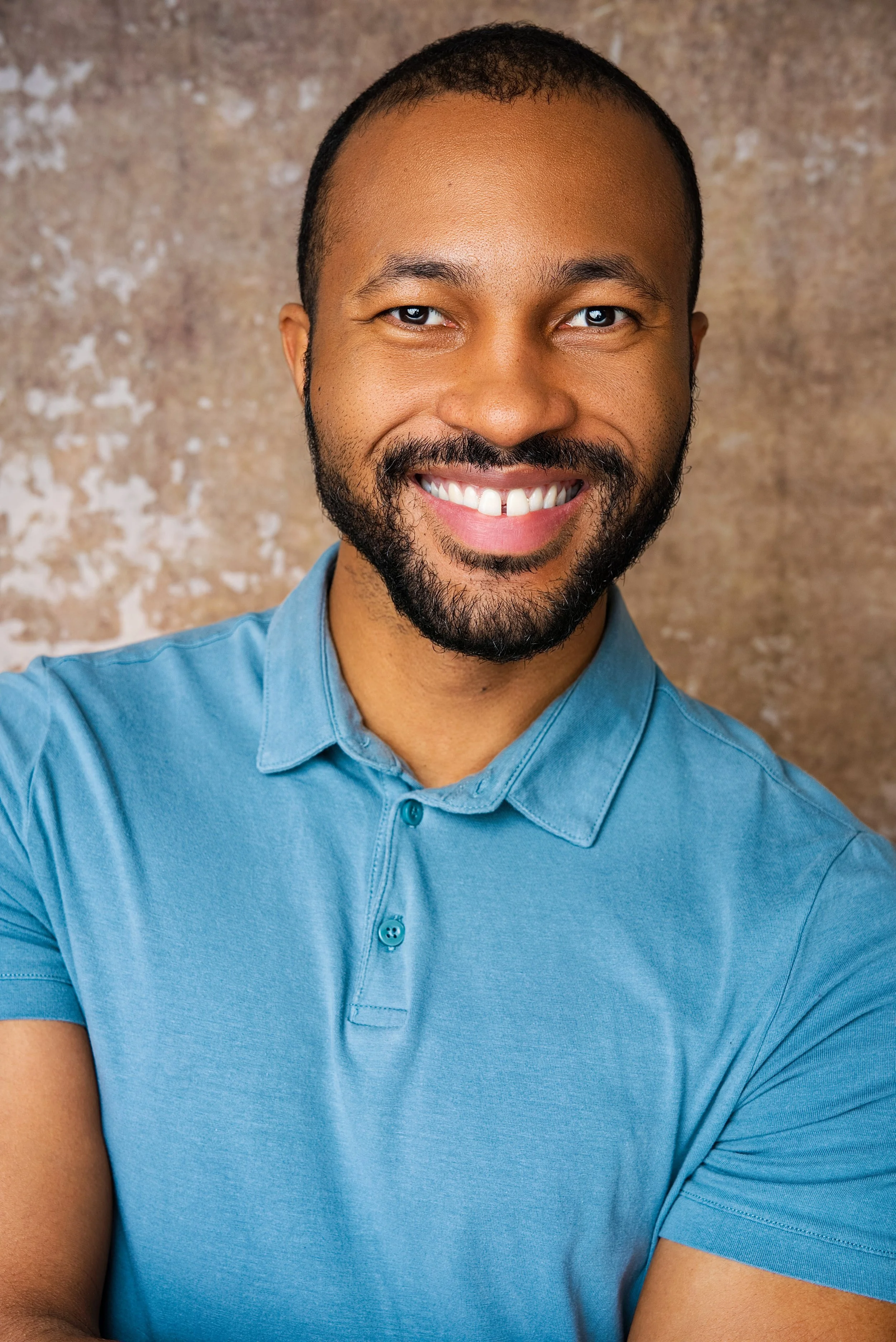 A man with a beard smiling, wearing a blue polo shirt, standing against a textured brown background.