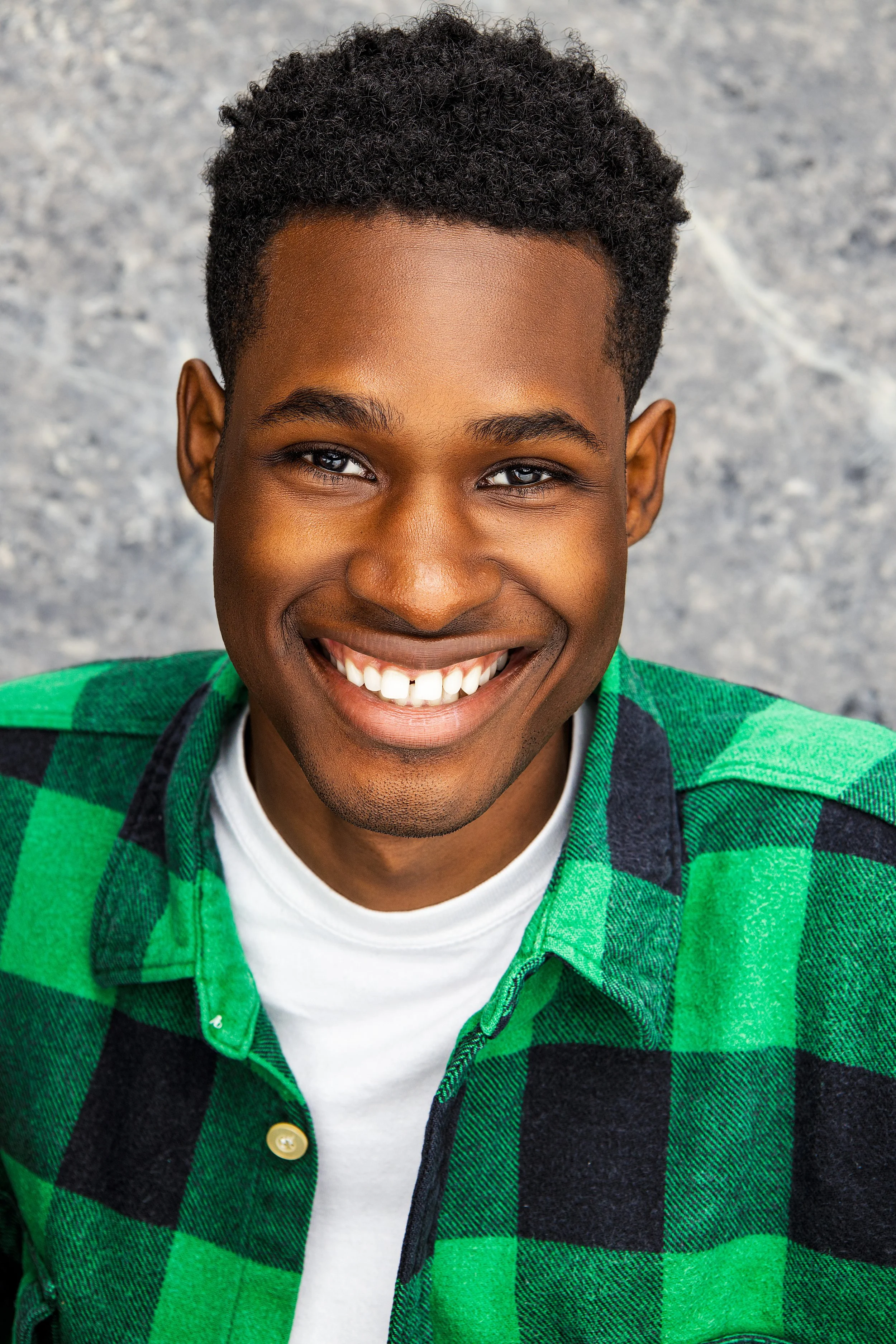 A smiling young man with dark curly hair wearing a green and black plaid shirt over a white t-shirt, standing against a gray background.