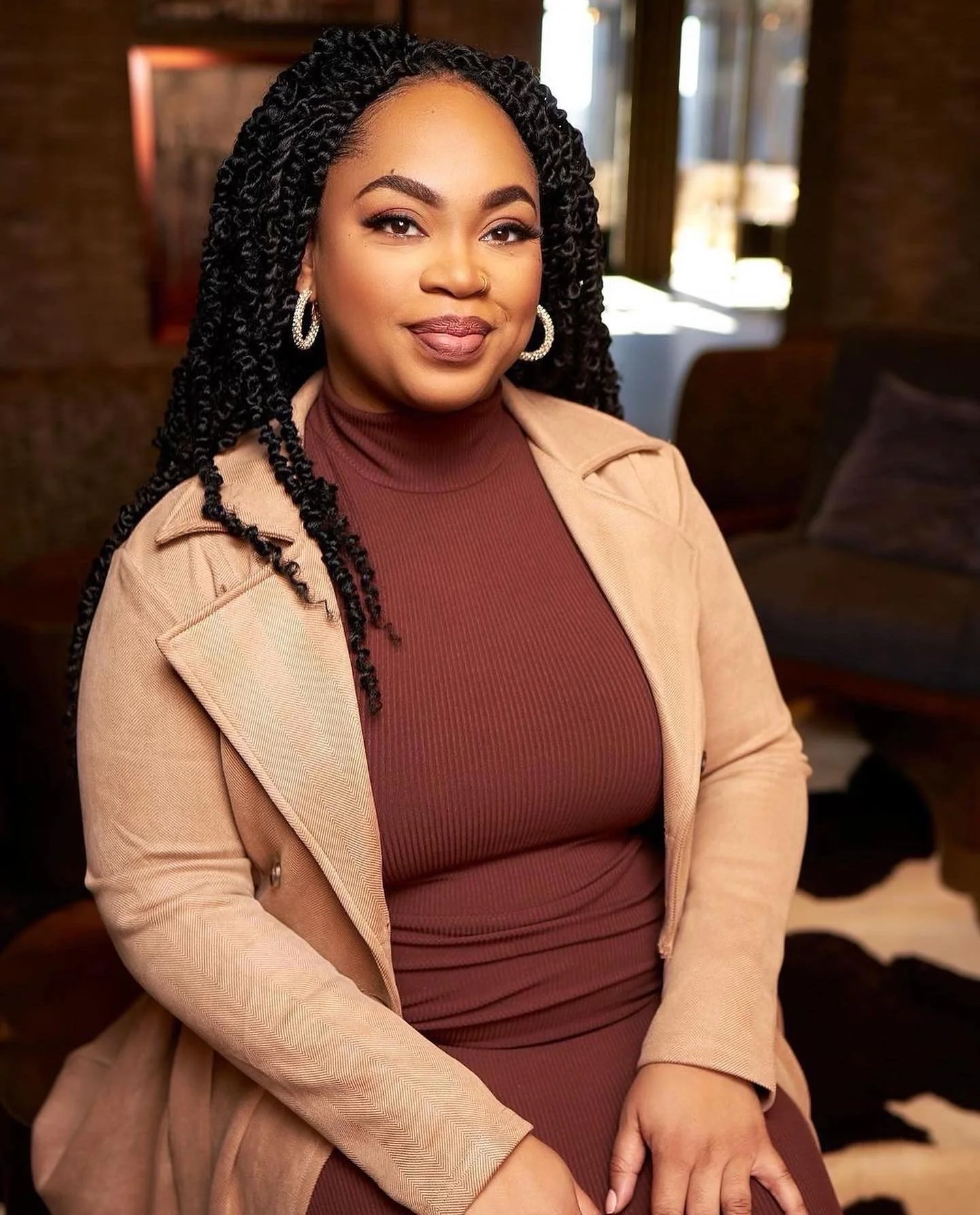 A woman with long, twisted black hair, wearing hoop earrings, a burgundy turtleneck, and a tan blazer, sitting indoors near a window with a couch in the background.