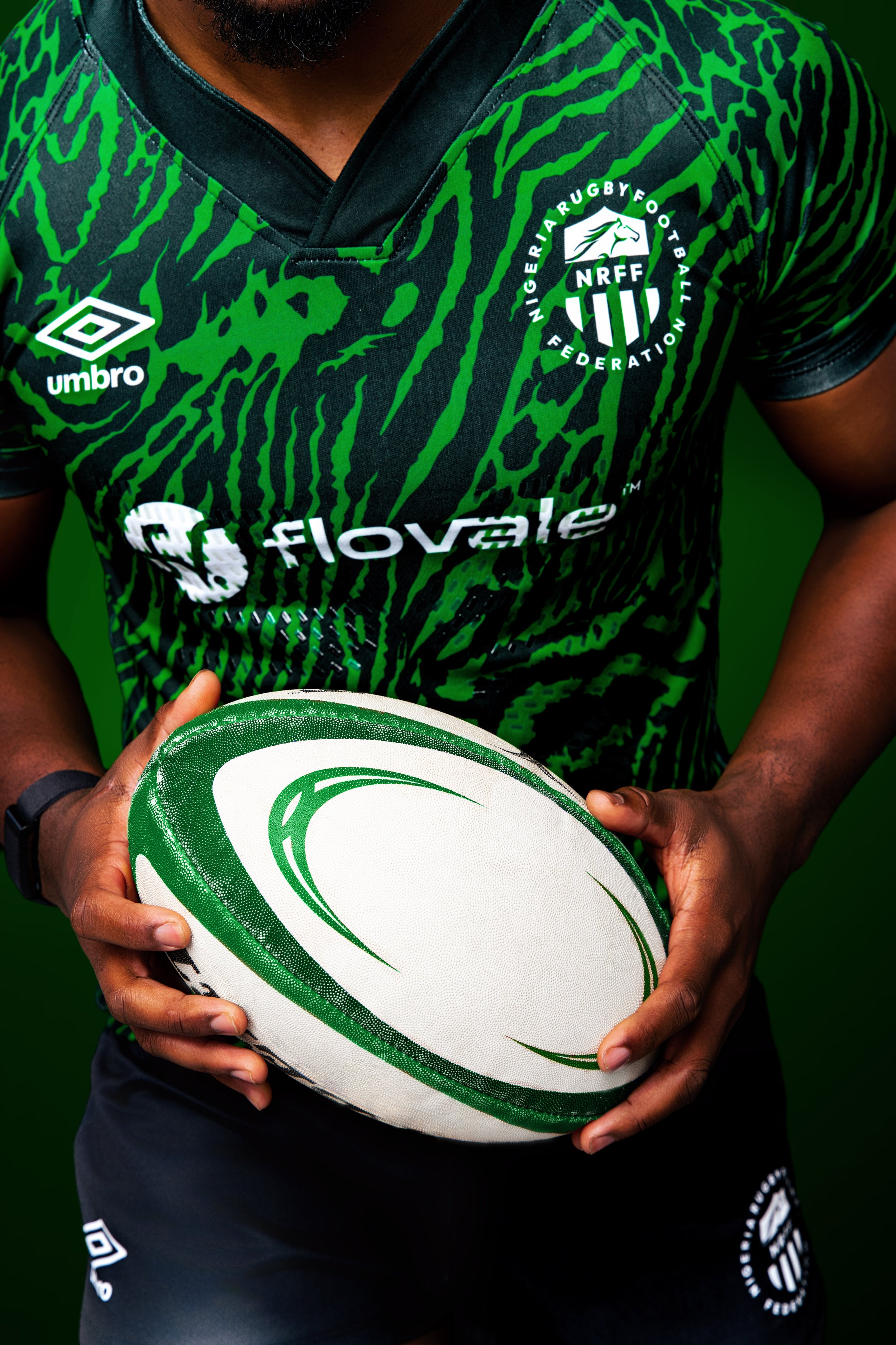 A rugby player wearing a black and green Nigeria Rugby Football Federation jersey, holding a rugby ball with green and white design, against a green background.
