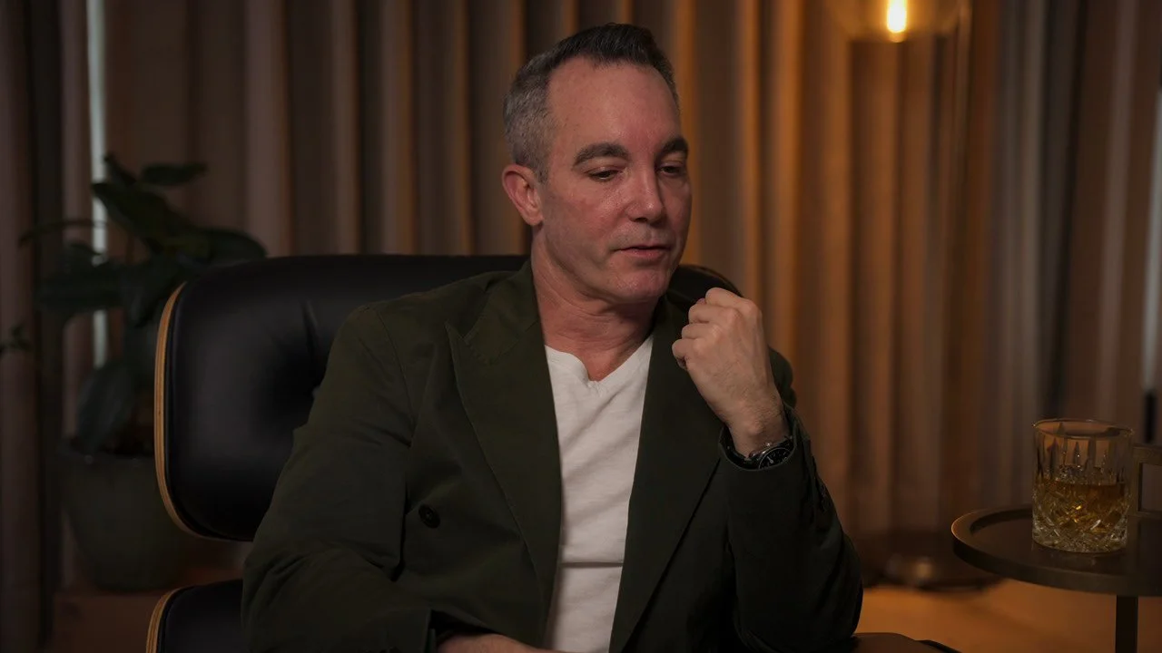 A man sitting in a black leather chair with a dark green blazer, white shirt, and a watch, in a room with brown curtains and a glass of brown liquid on a side table.