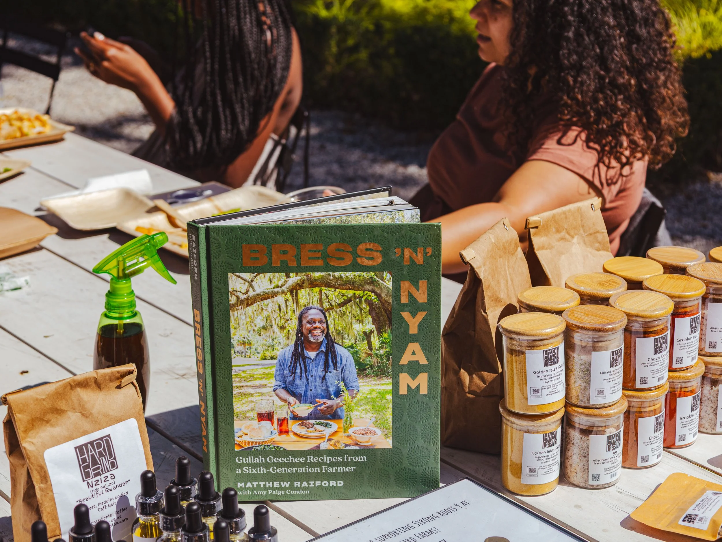 A cookbook titled 'BRESS 'N NYAM' featuring a photo of a smiling man with dreadlocks holding a plate outdoors, surrounded by jars of spices and other food items on a rustic table at an outdoor market or event.
