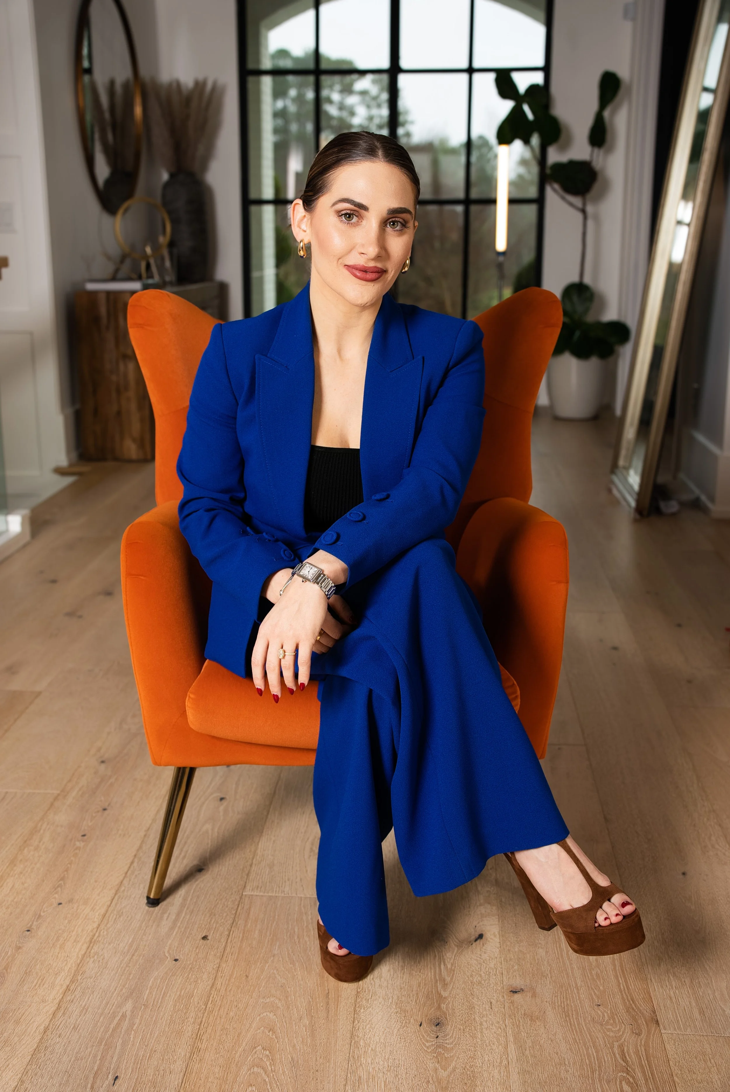 A woman with dark hair pulled back, wearing gold earrings and a royal blue suit, sits confidently in an orange armchair inside a modern home with large windows and hardwood floors.