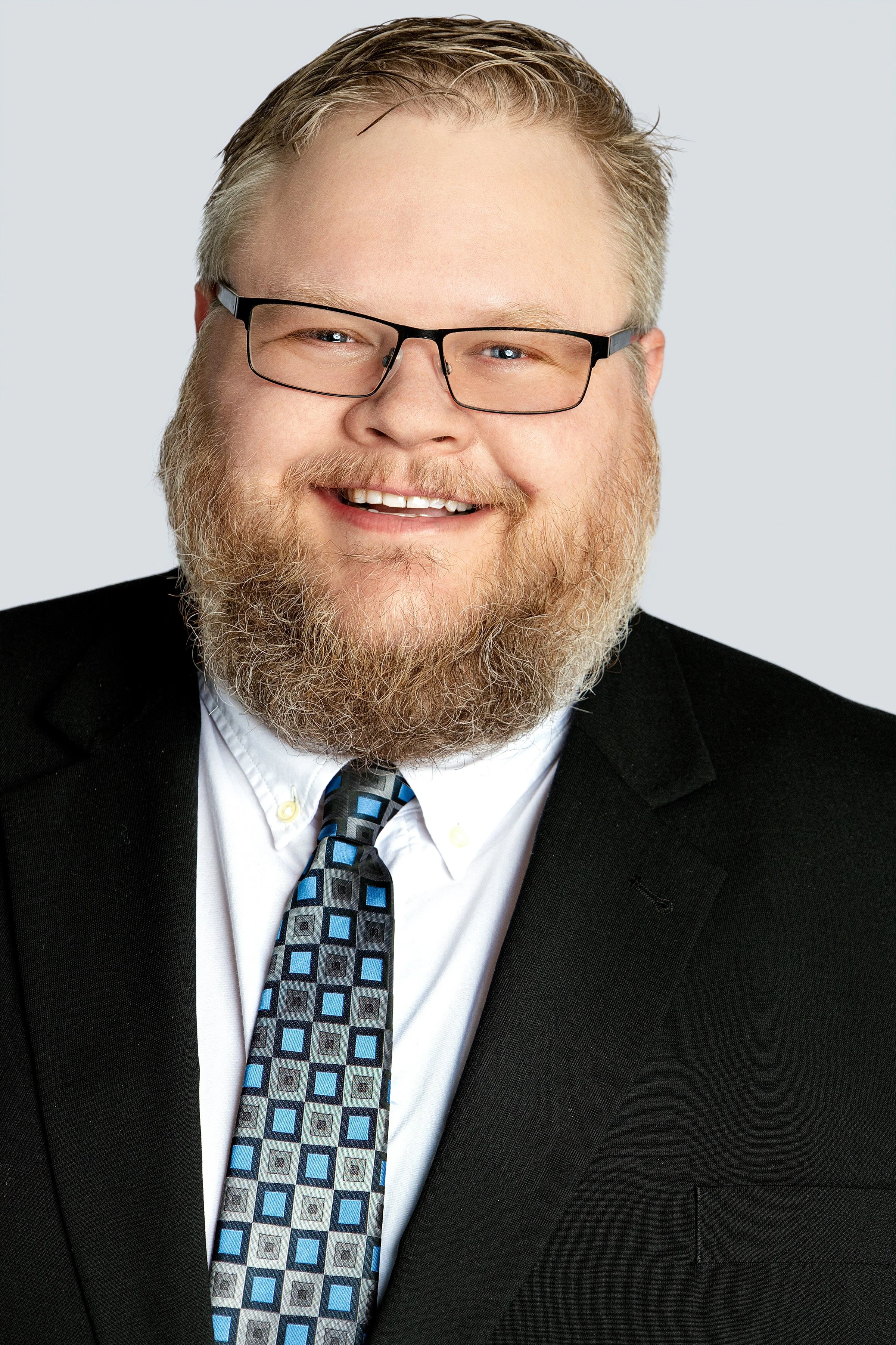 Portrait of a man with blond hair, glasses, a beard, wearing a black suit, white shirt, and a patterned tie, smiling against a plain background.
