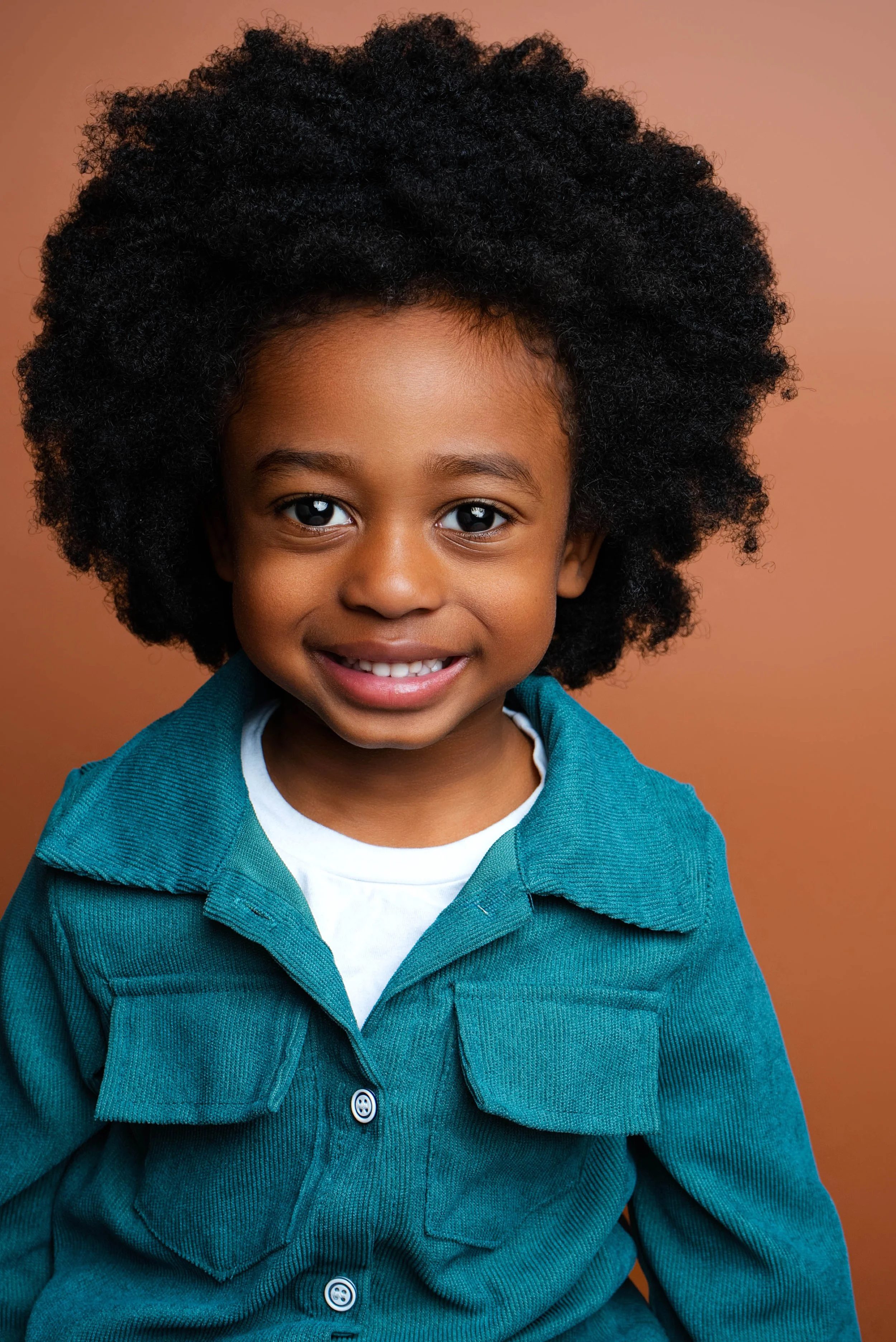 A young girl with curly hair wearing a teal corduroy jacket and white shirt, smiling at the camera against a light brown background.