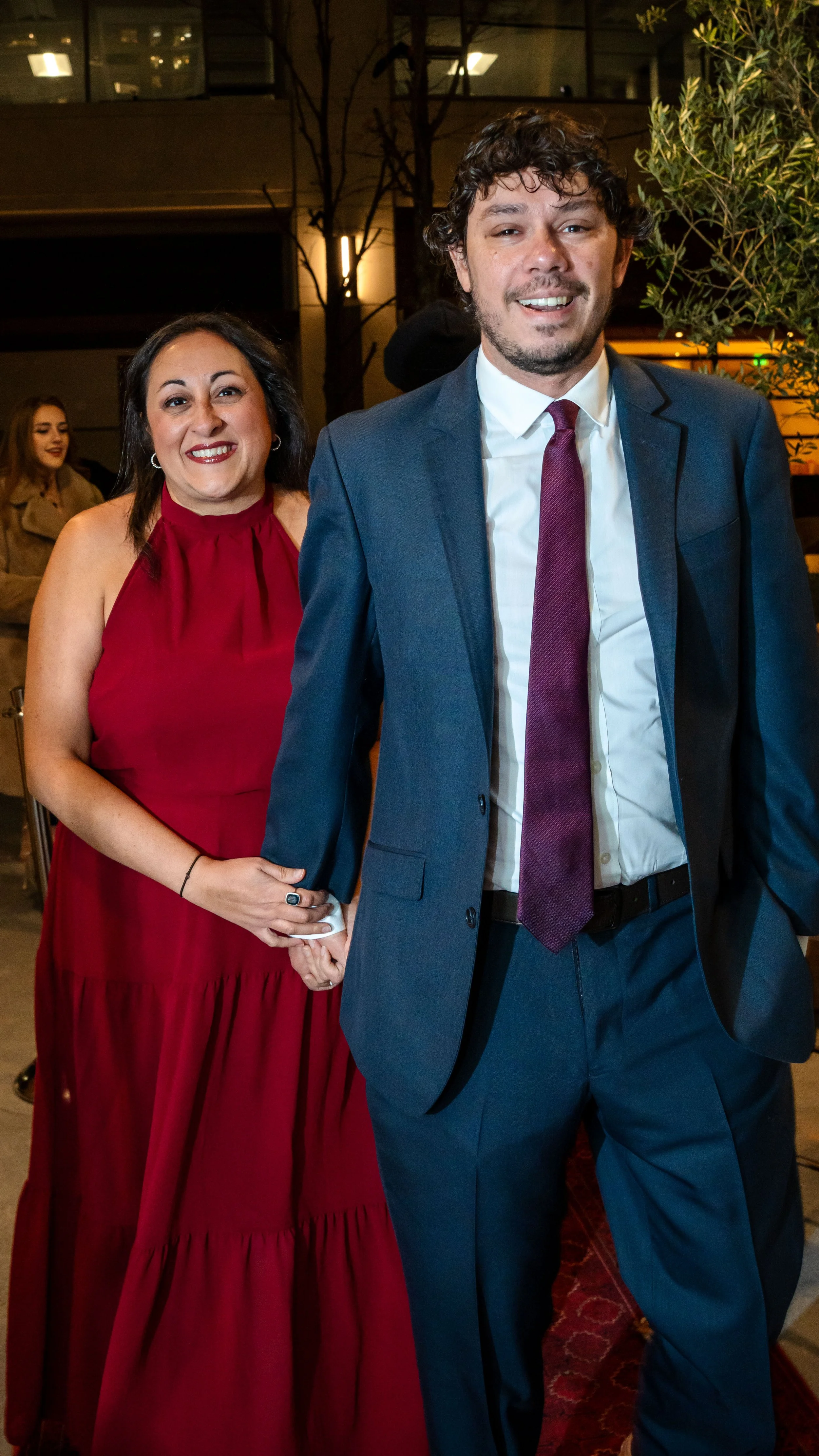 A man in a blue suit and white shirt holding a woman's hand, with a woman in a red dress smiling behind him at an indoor event.