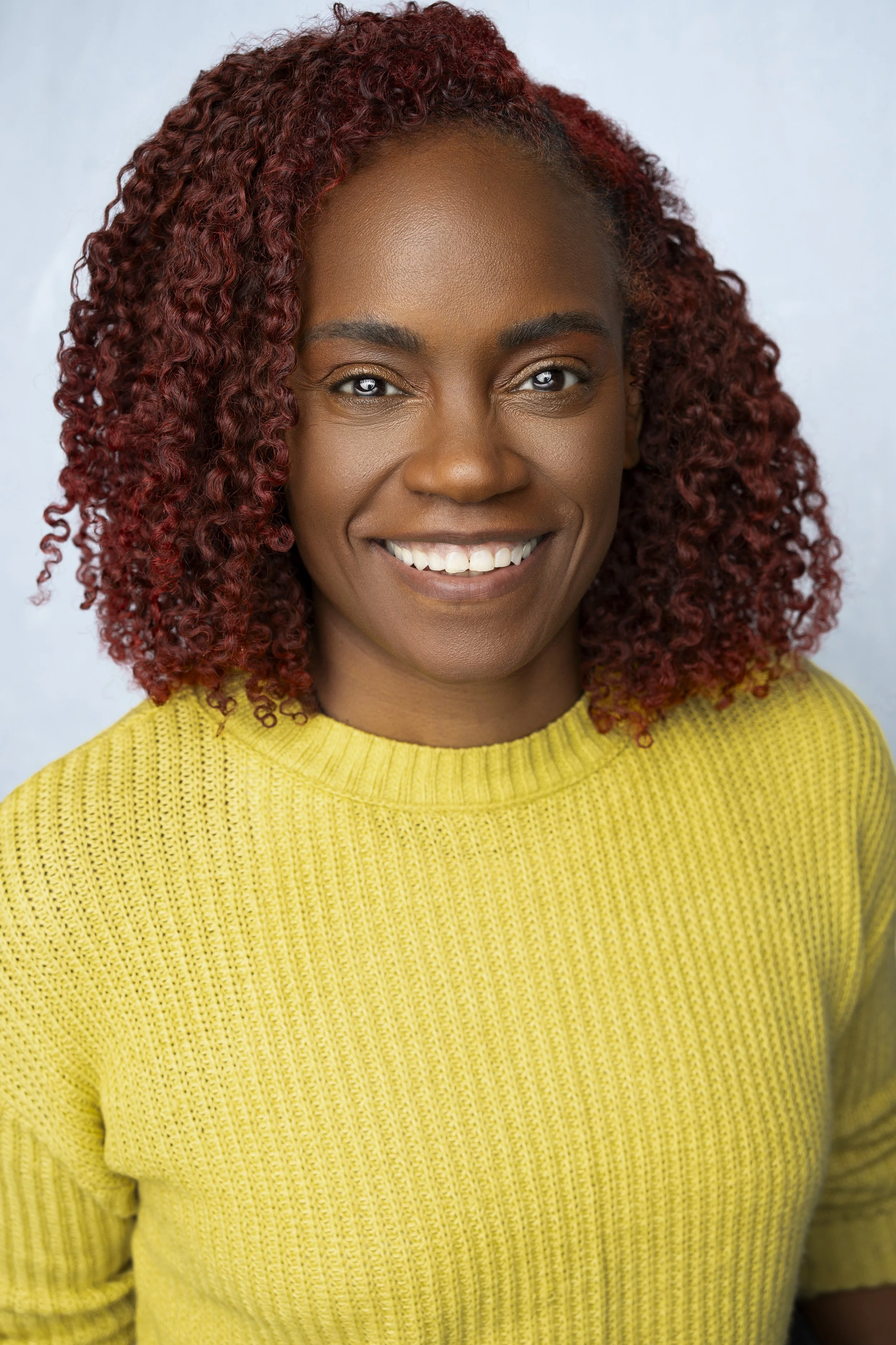 Portrait of a woman with curly red hair, smiling, wearing a yellow knit sweater, against a light background.