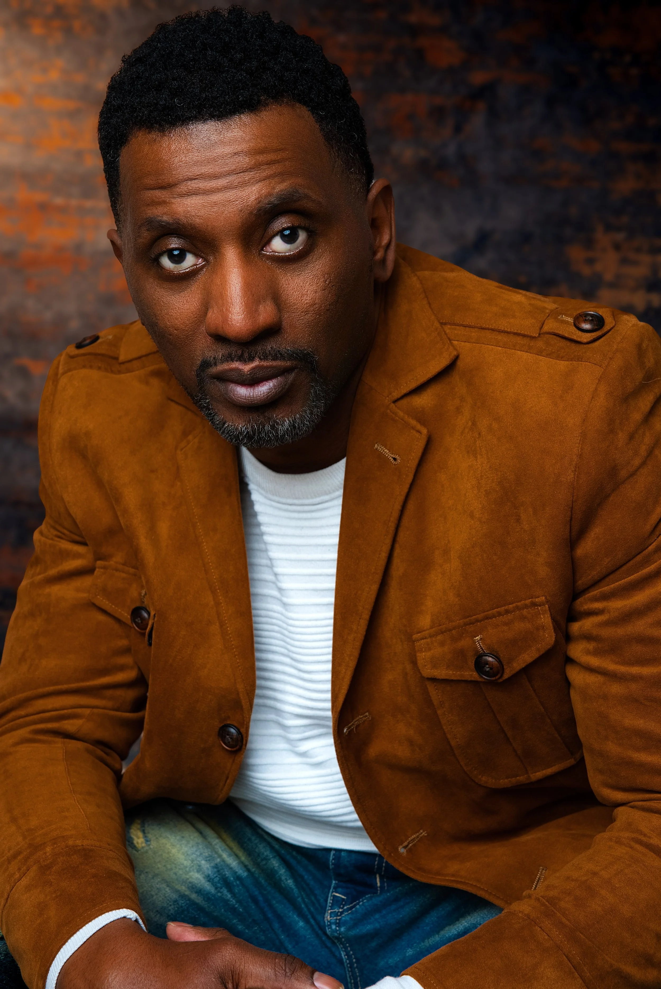 A man with short curly black hair and a beard in a brown jacket and white shirt, sitting against a dark, textured background.