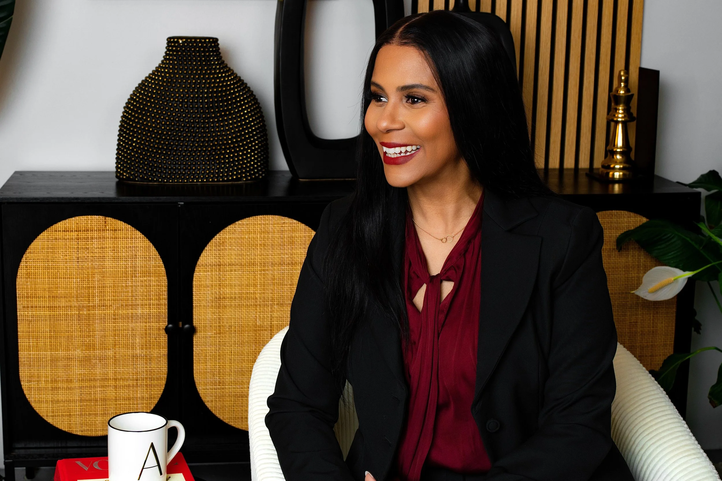 A woman with long dark hair, wearing a black blazer and a red blouse, smiling while sitting in a modern room with decorative items and a white mug on a red book nearby.