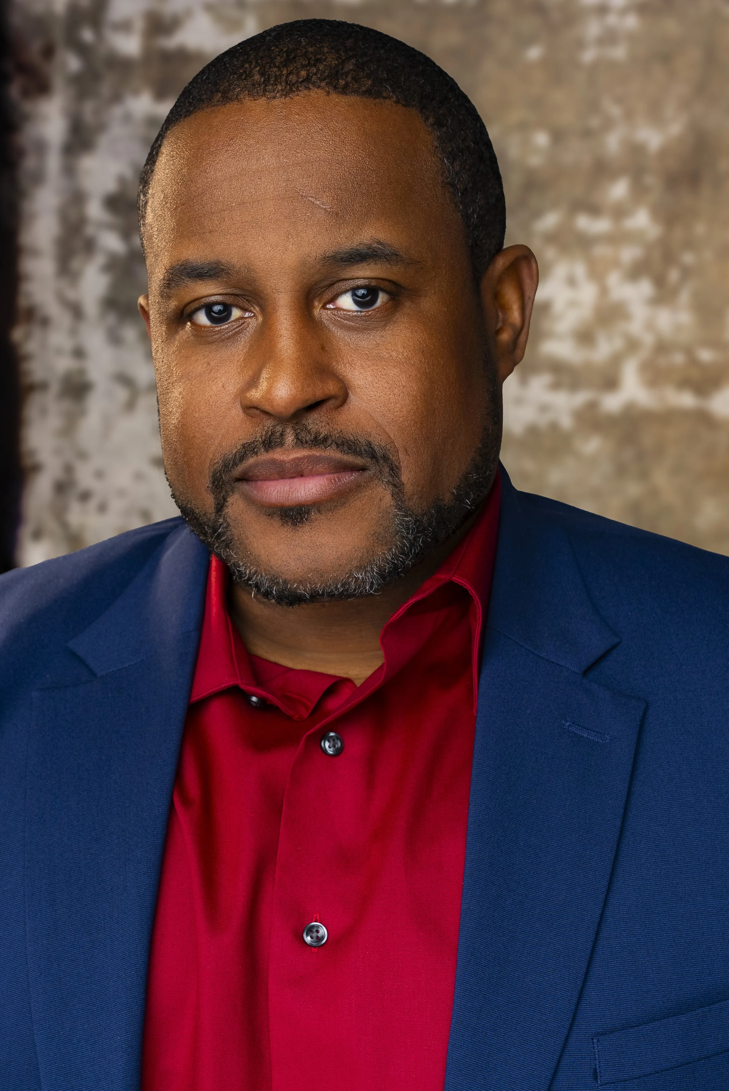 A professional headshot of a man wearing a red shirt and a blue blazer, with a textured brown background.