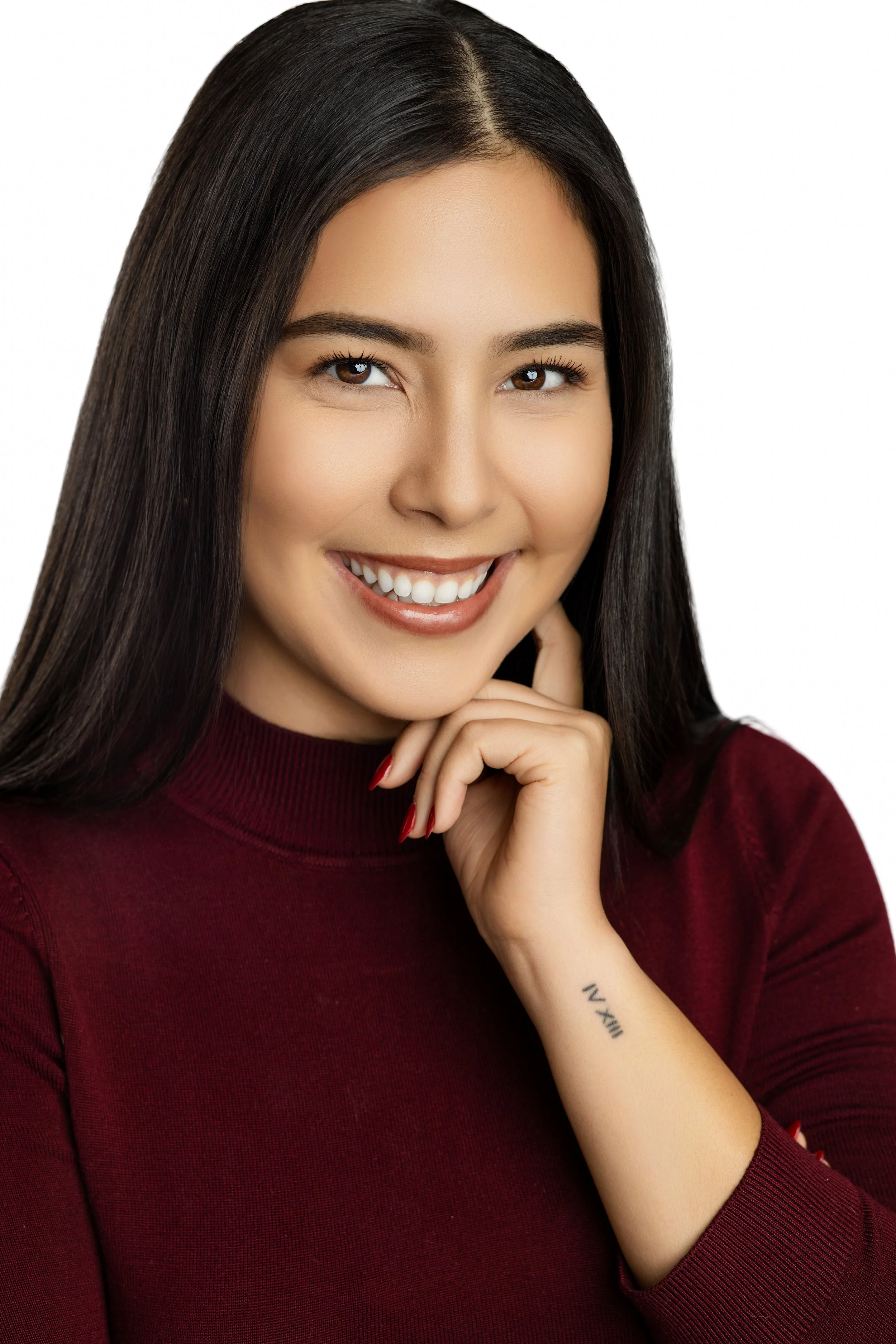 A smiling woman with long dark hair, wearing a burgundy long sleeve top, with one hand resting near her chin, and a tattoo on her wrist that says 'XVI'.