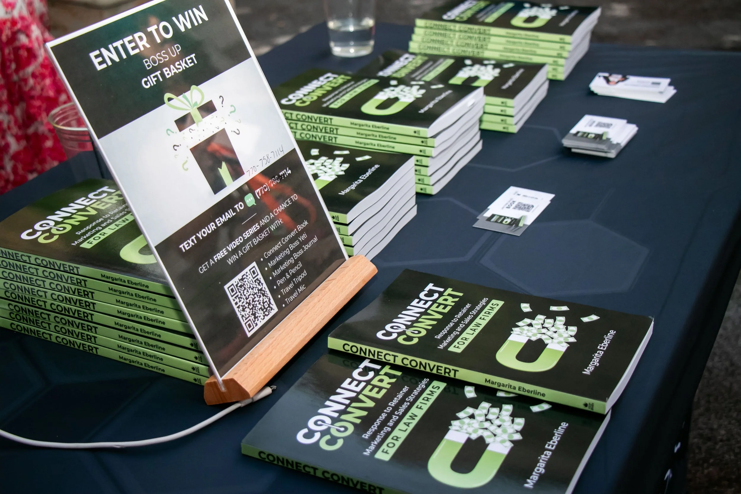 Table displaying copies of the book 'Connect Convert' by Margarita Eberline, promotional flyers, a sign offering a gift basket raffle, and small stacks of business cards at an event.